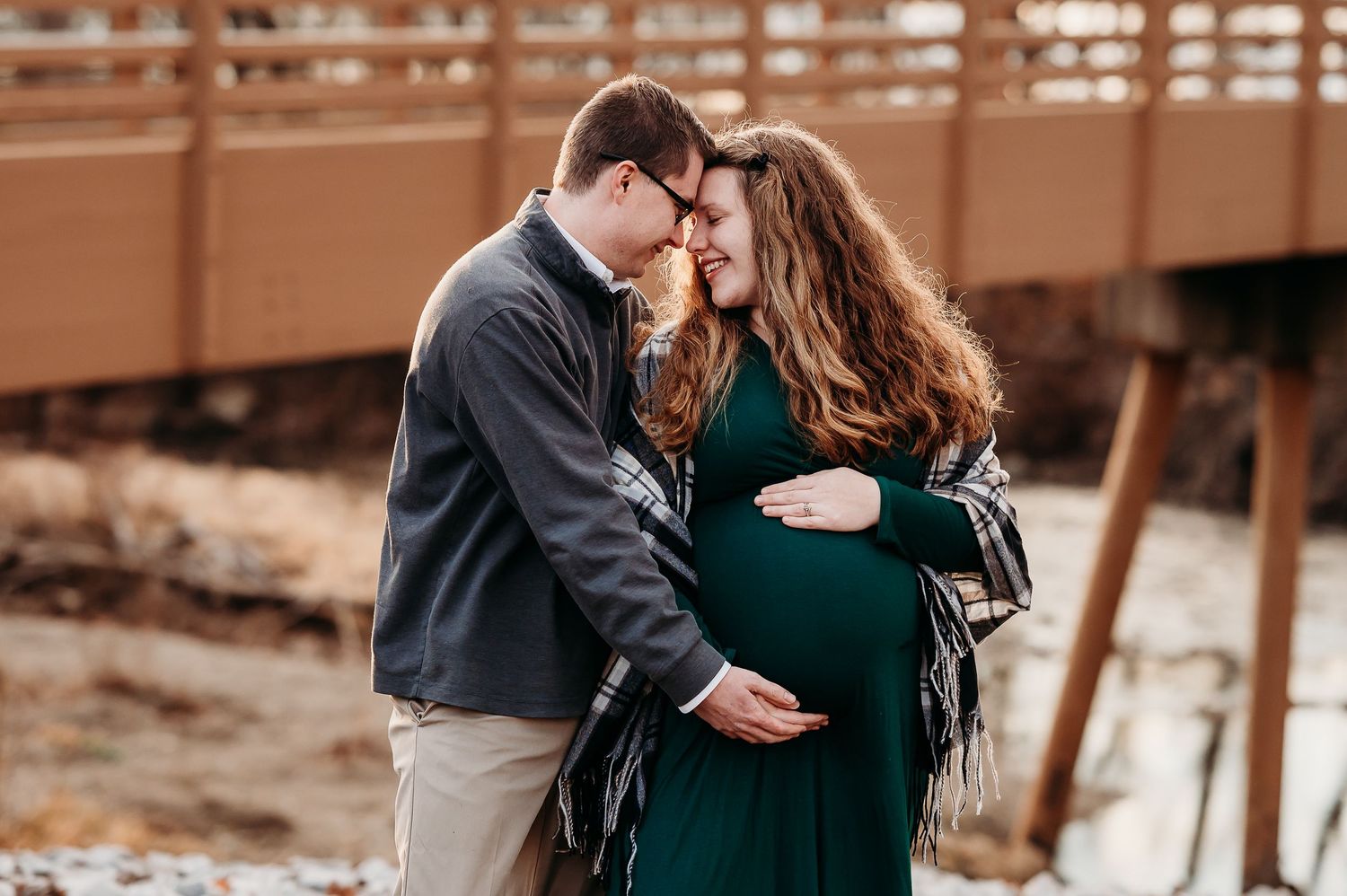 Couple sharing tender moment during Plainfield outdoor maternity photoshoot near wooden bridge, woman in green dress with plaid wrap.