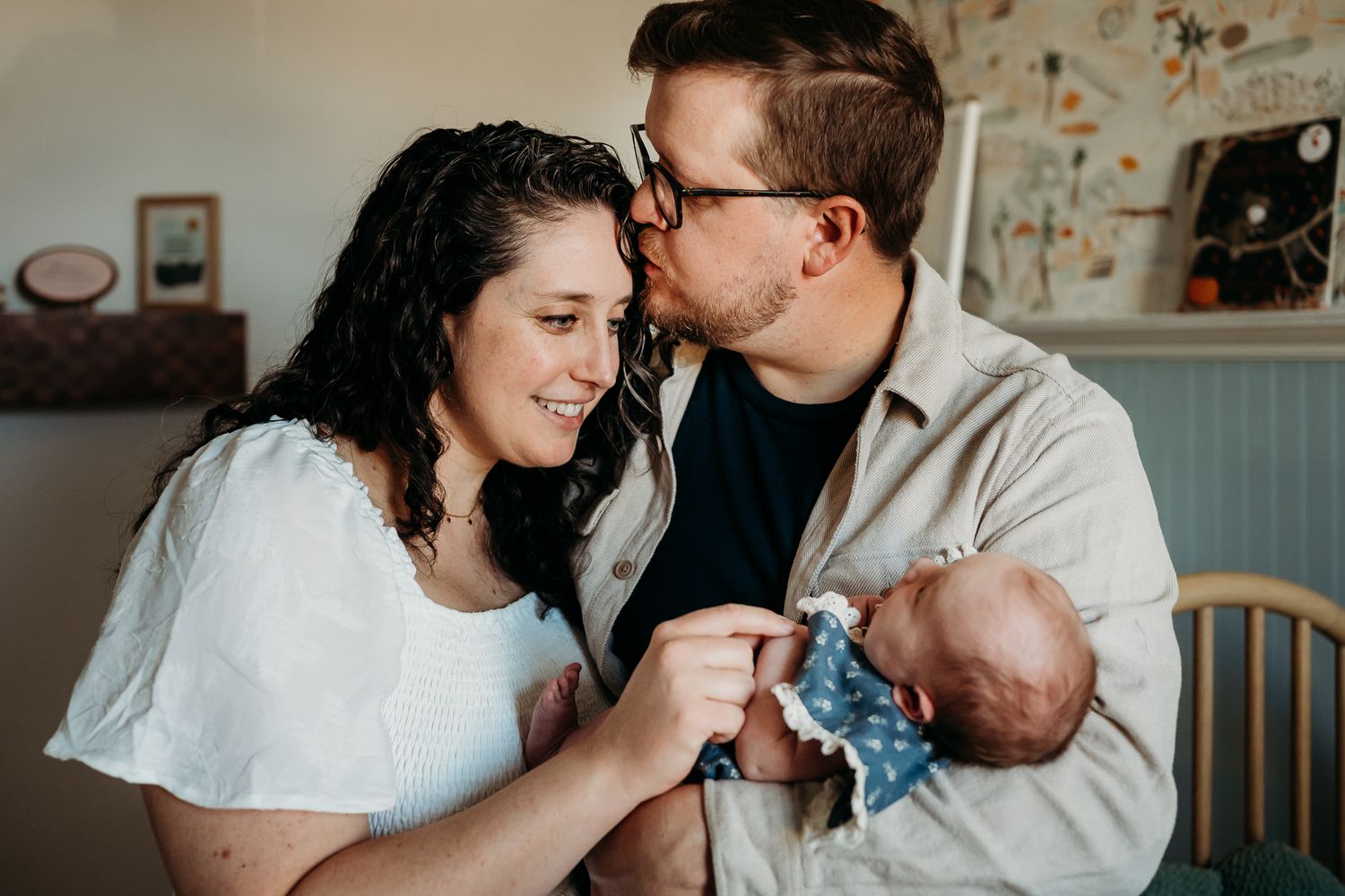 Husband kisses wife while holding their baby girl during lifestyle newborn photo session in their Westfield, Indiana, home nursery.