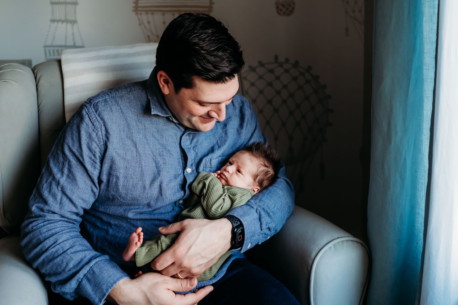 A person in a blue shirt sits in a cushioned chair tenderly holding a sleeping newborn baby wrapped in blankets.