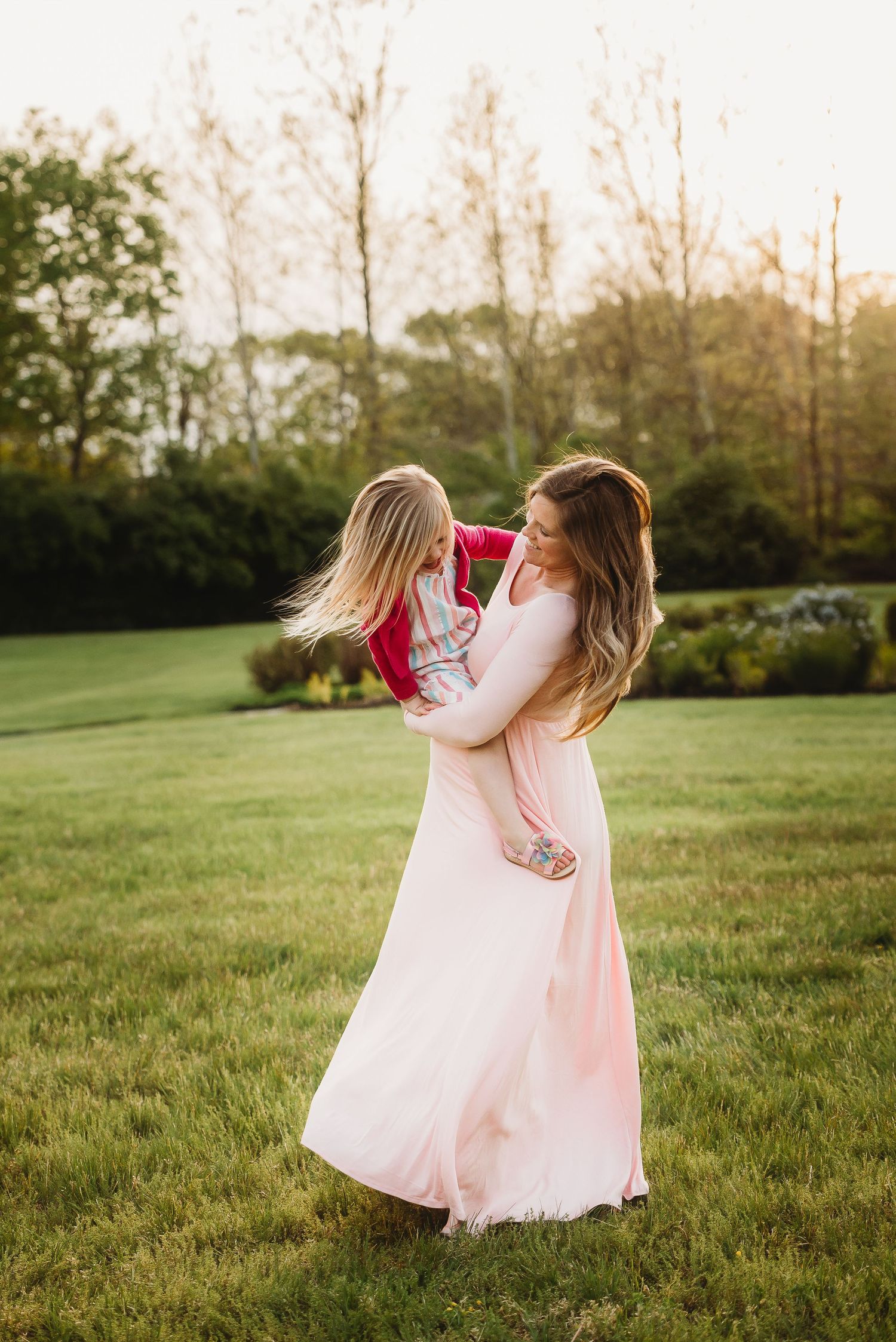 Mom and little girl in pink dresses share a joyful moment spinning together at sunset during spring maternity photos in Plainfield, Indiana.