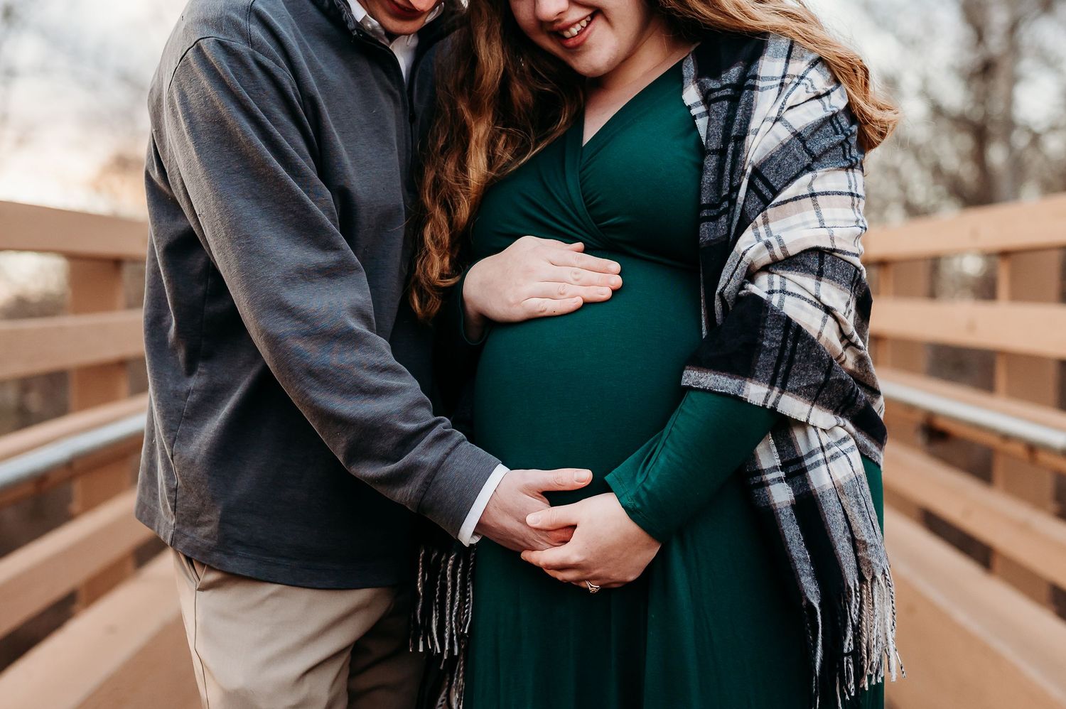 Couple cradling pregnant belly during maternity photo shoot on wooden bridge, woman in green dress with plaid shawl.