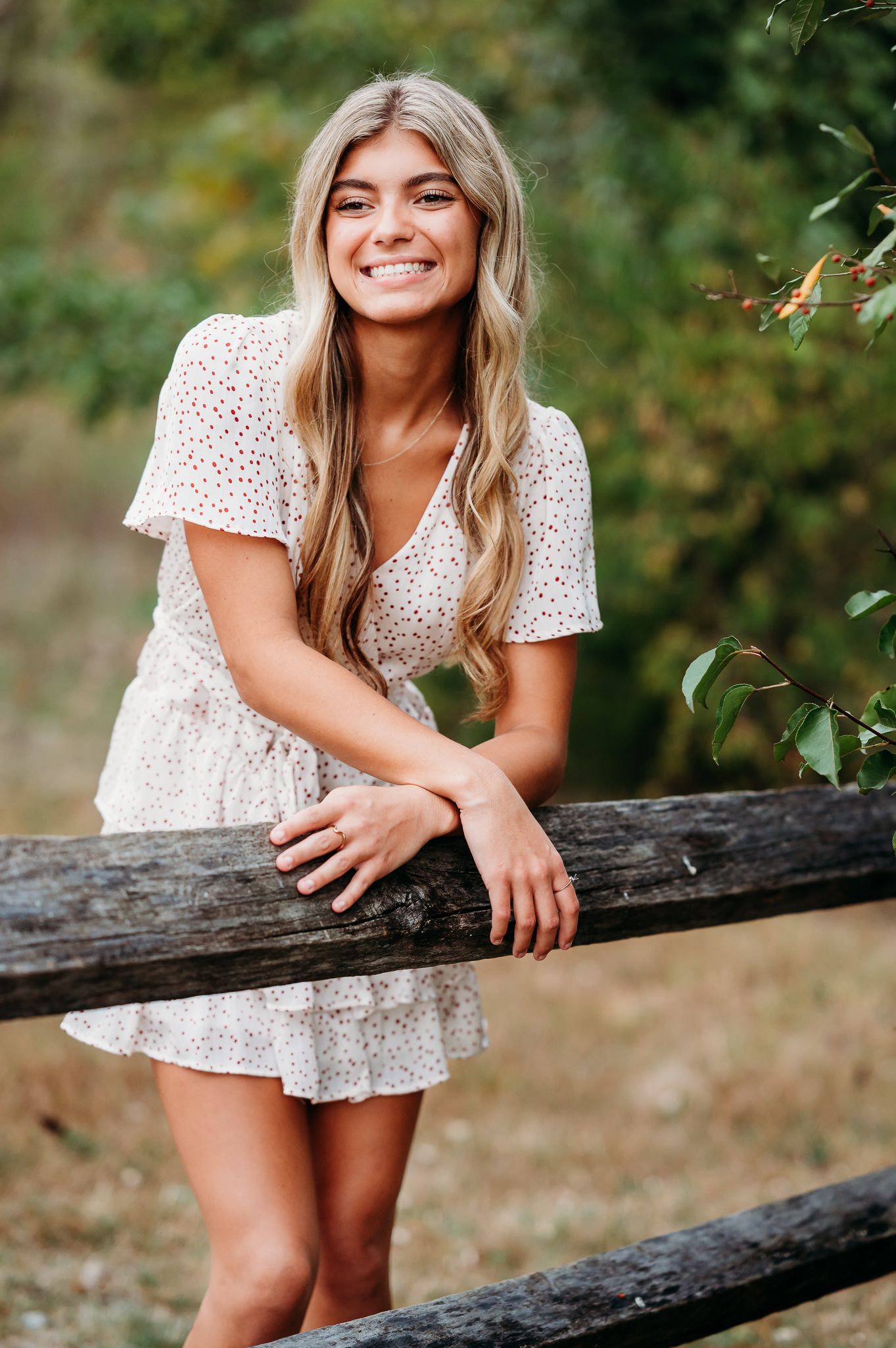 A person in a white polka dot dress leans casually against a wooden fence in an outdoor setting with greenery.