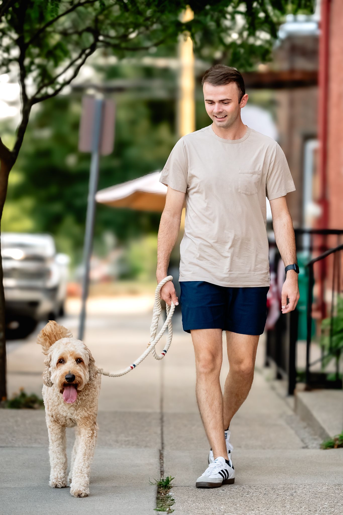 Young man in beige t-shirt and blue shorts walks fluffy goldendoodle on leash along Danville sidewalk during dog photoshoot with Sara House.