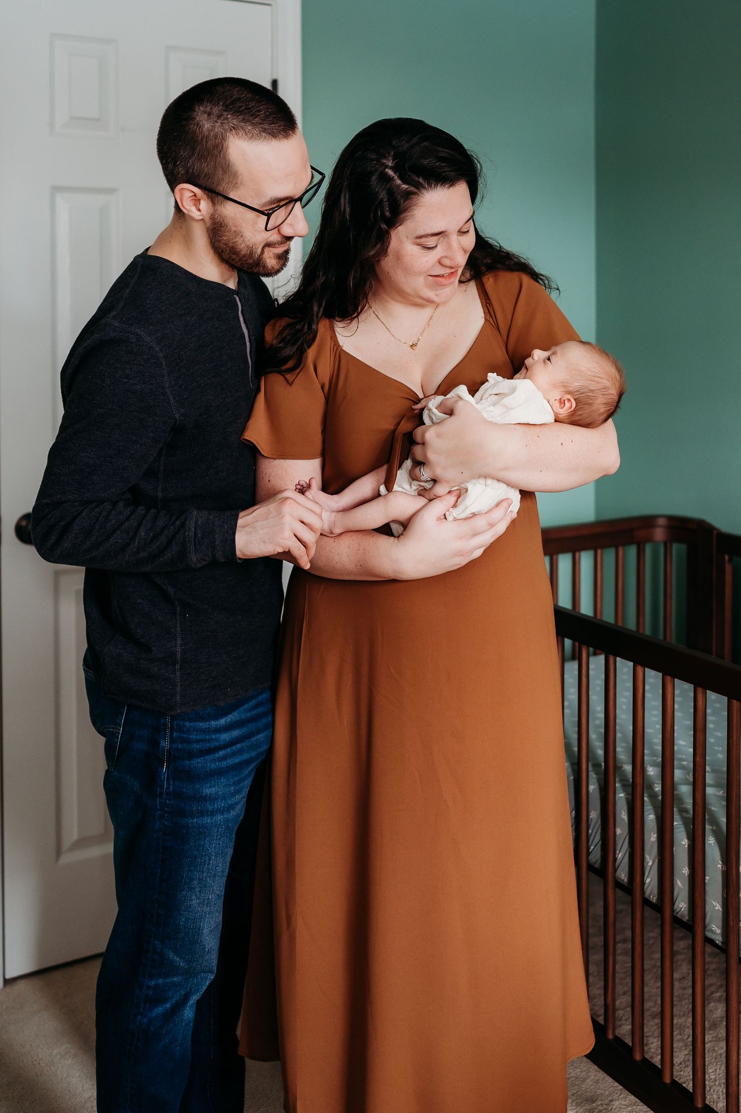 Noblesville, Indiana, parents stand lovingly together with their newborn baby in a nursery with a wooden crib.