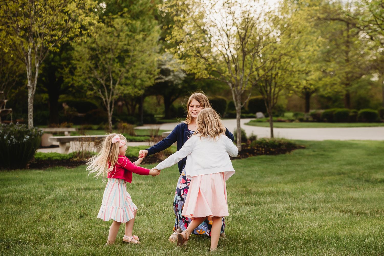 Three sisters hold hands while spinning together through a grassy outdoor area during golden hour photoshoot in Friendship Gardens.