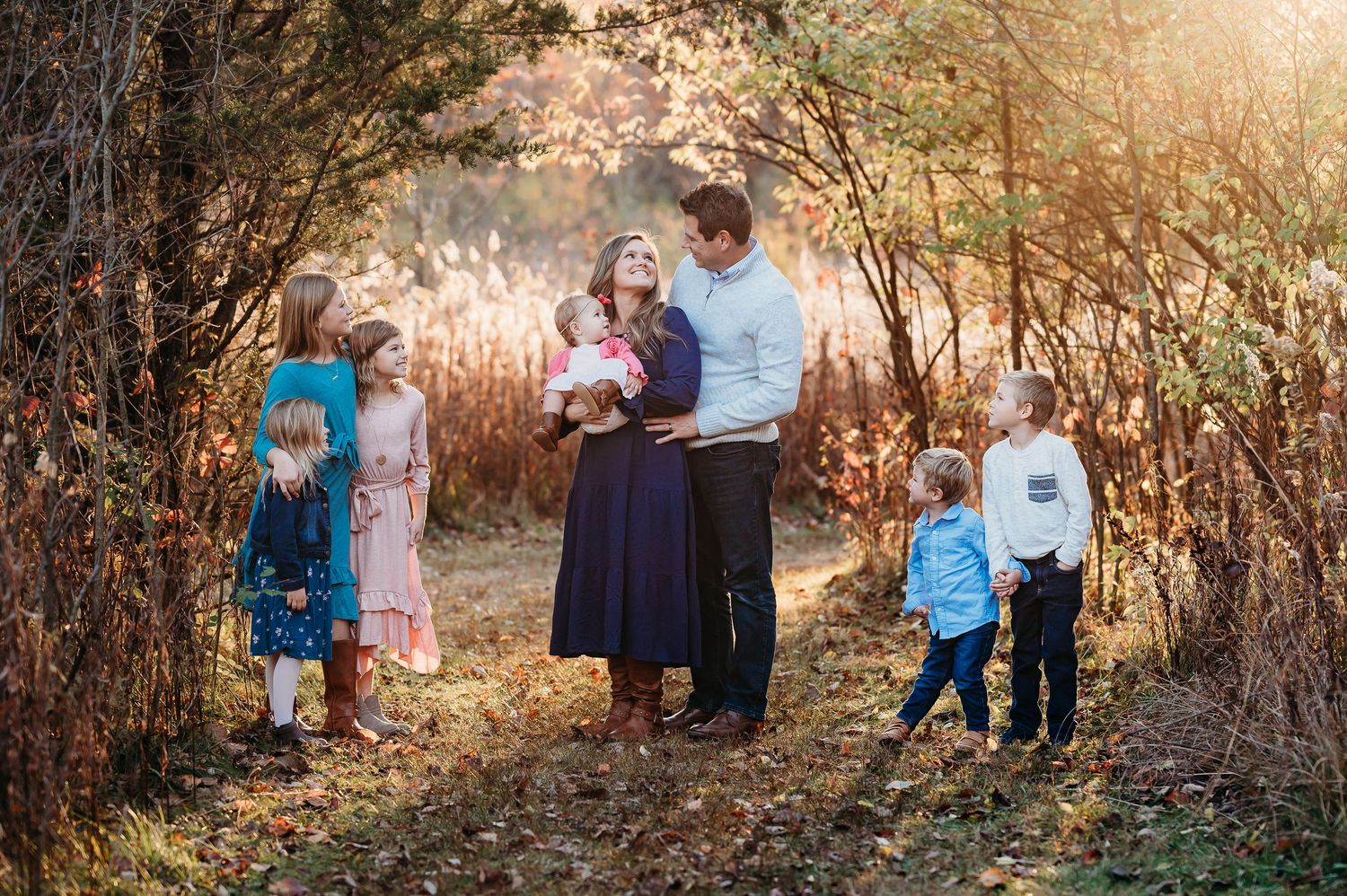 A family stands together on wooded path during golden hour with sunlight filtering through autumn trees at Ritchey Woods in Fishers, Indiana