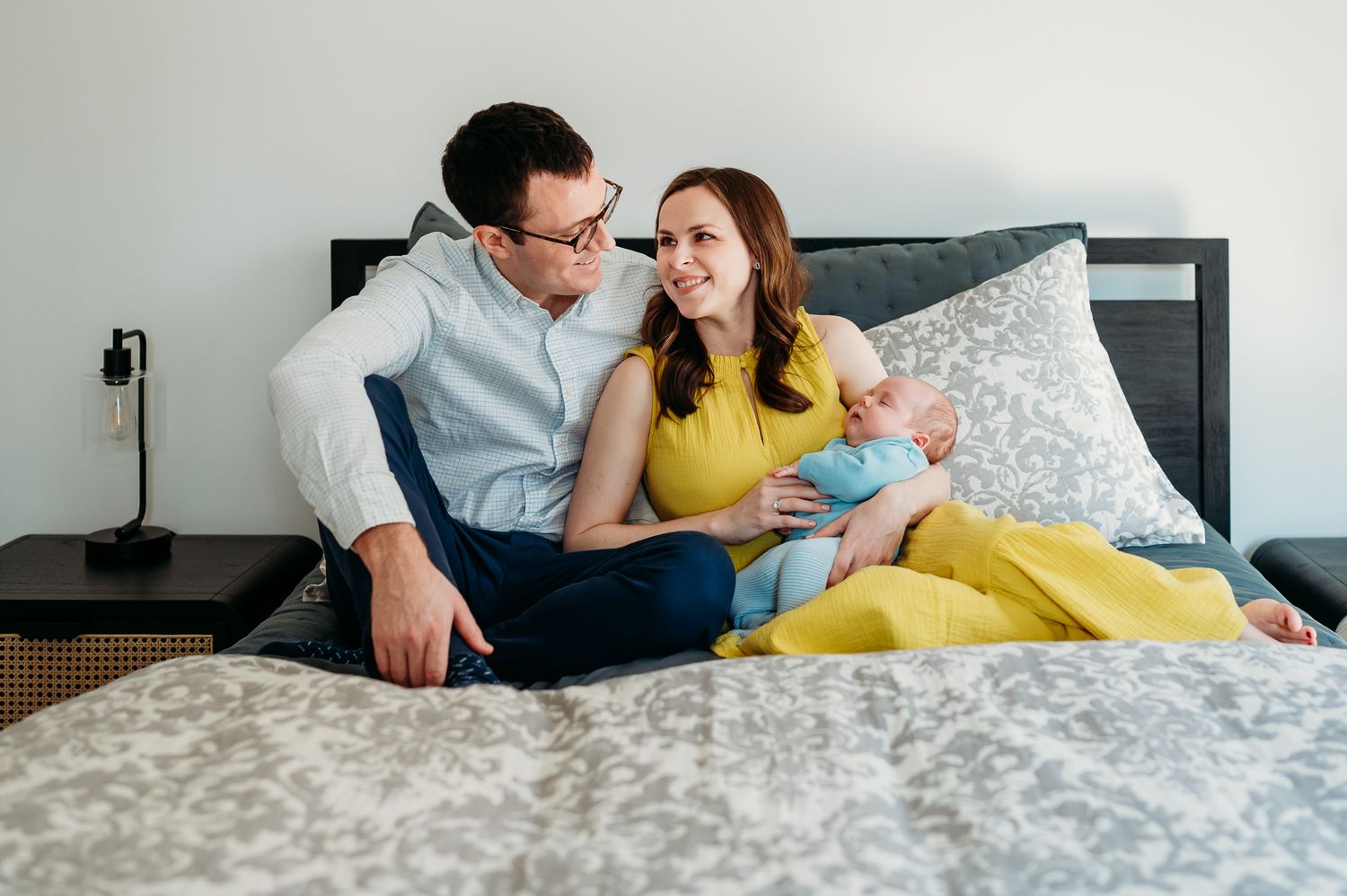 A couple relaxes on a gray bed while holding their infant child, with mom in a yellow dress during Westfield lifestyle newborn photo session