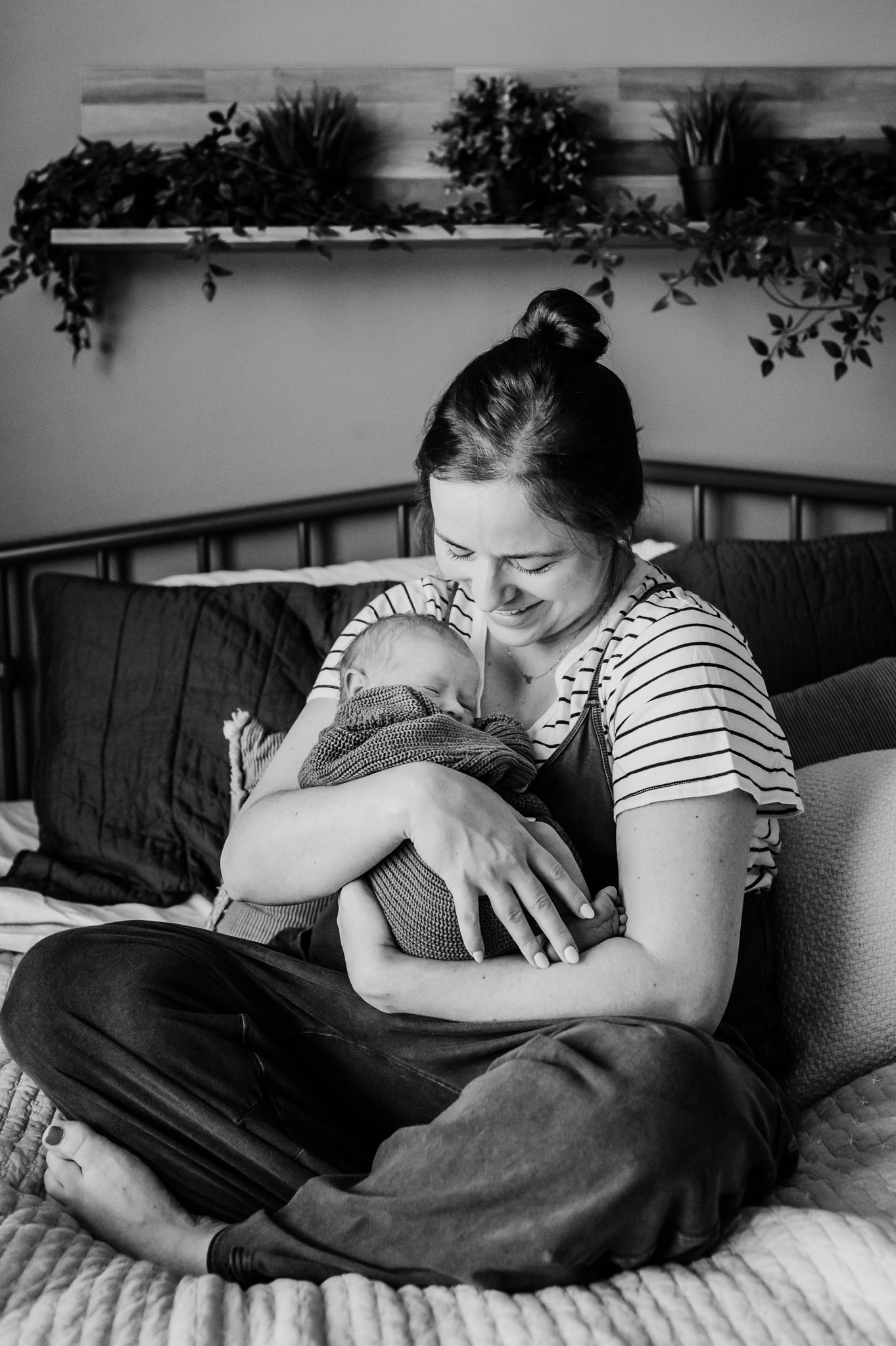 A tender nurturing moment captured in black and white as a mother cradles her newborn baby on a cozy bed in Indiana home.
