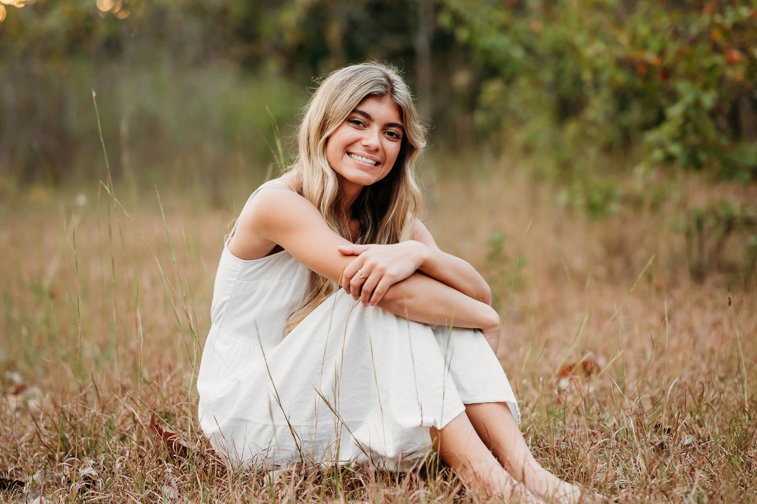 Someone in a white dress sits on dry grass in a peaceful outdoor setting during golden hour.