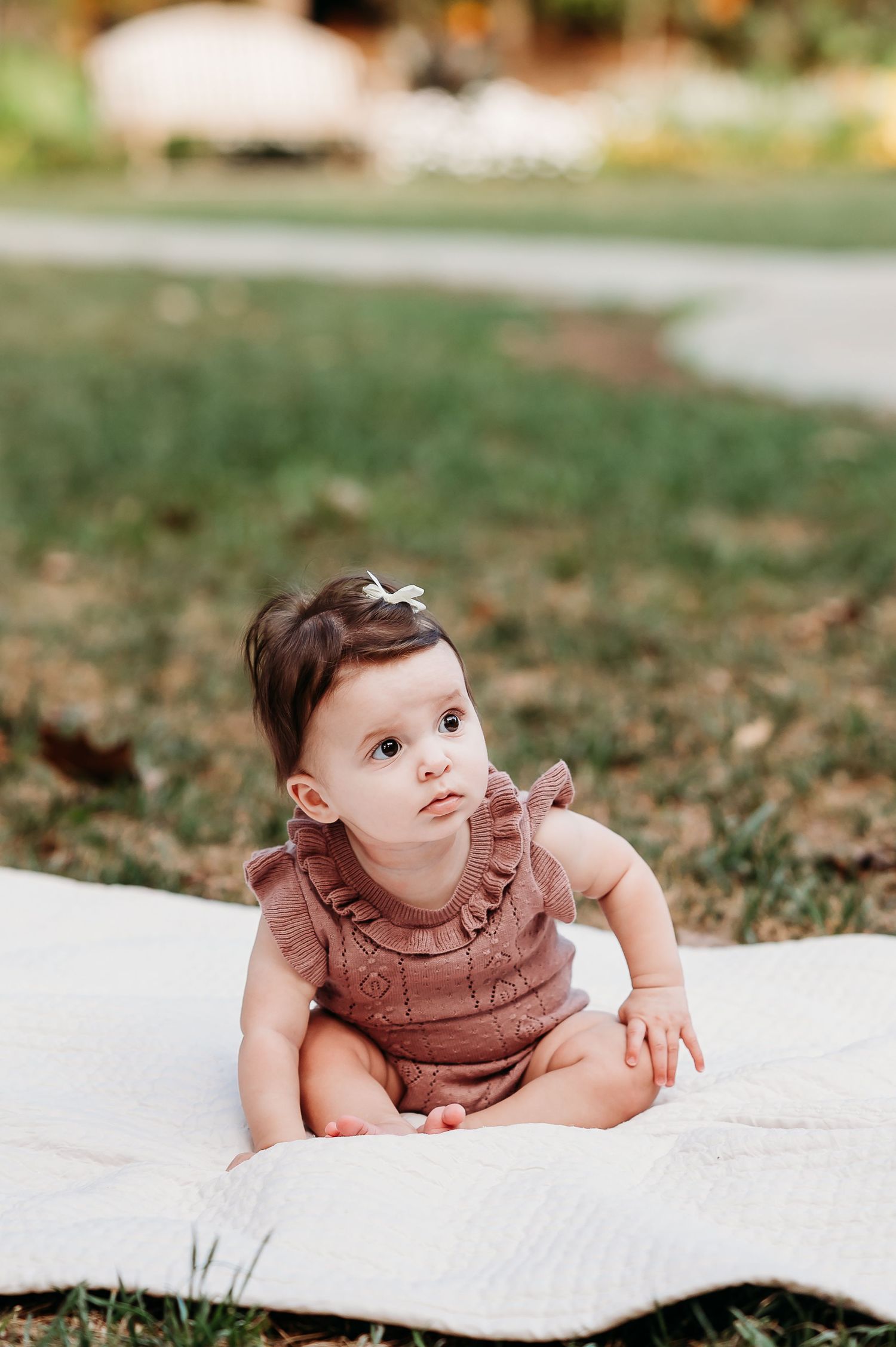 Baby girl in pink ruffle outfit sitting on white blanket outdoors during summer photoshoot at Newfields in Indianapolis.