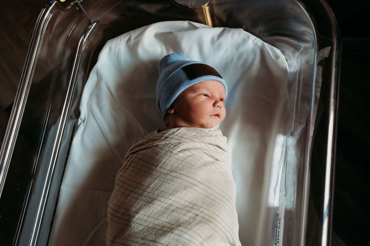 A newborn baby wearing a blue hat lies swaddled in a white blanket in a hospital bassinet.