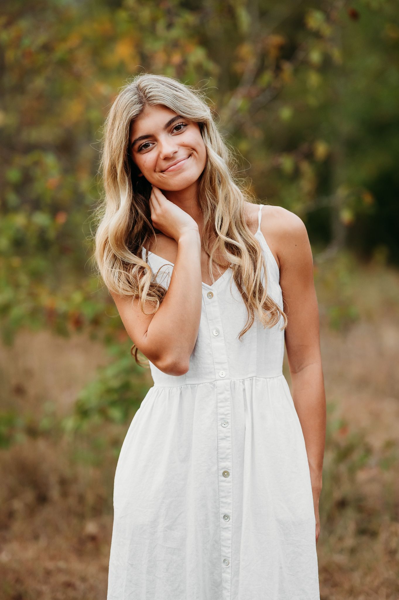 Portrait of someone in a white button-up sundress with long wavy blonde hair posing outdoors against a natural background.