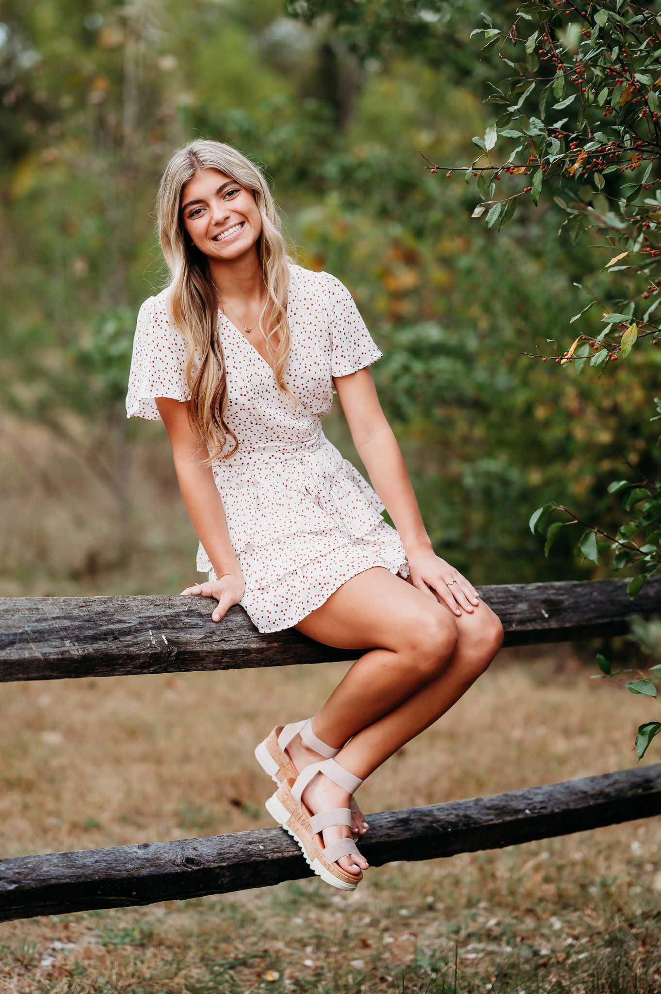 Someone in a white floral dress and sandals sits casually on a wooden fence surrounded by greenery on a sunny day.