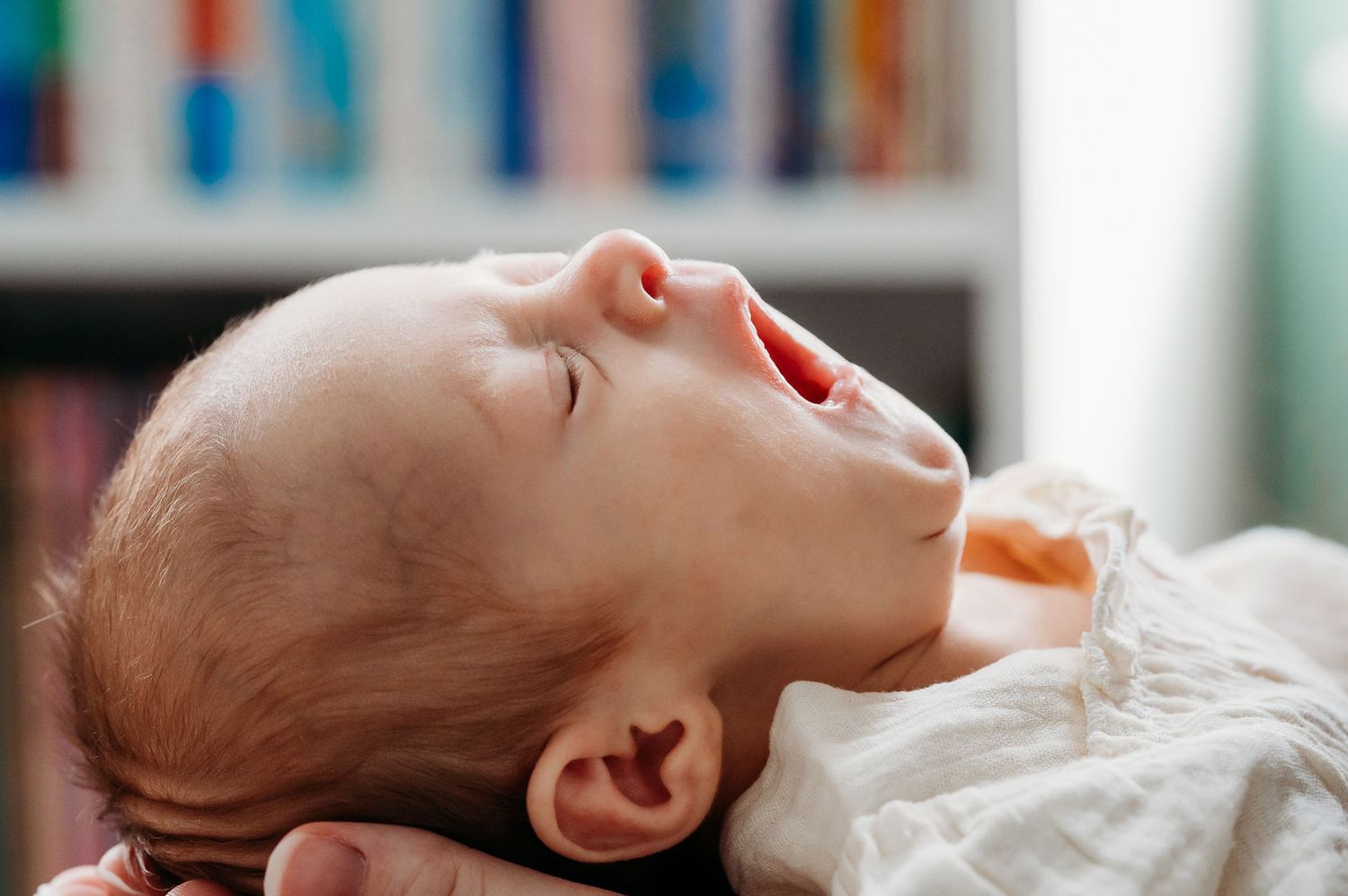 A baby wearing a white onesie rests on a soft surface in natural light.
