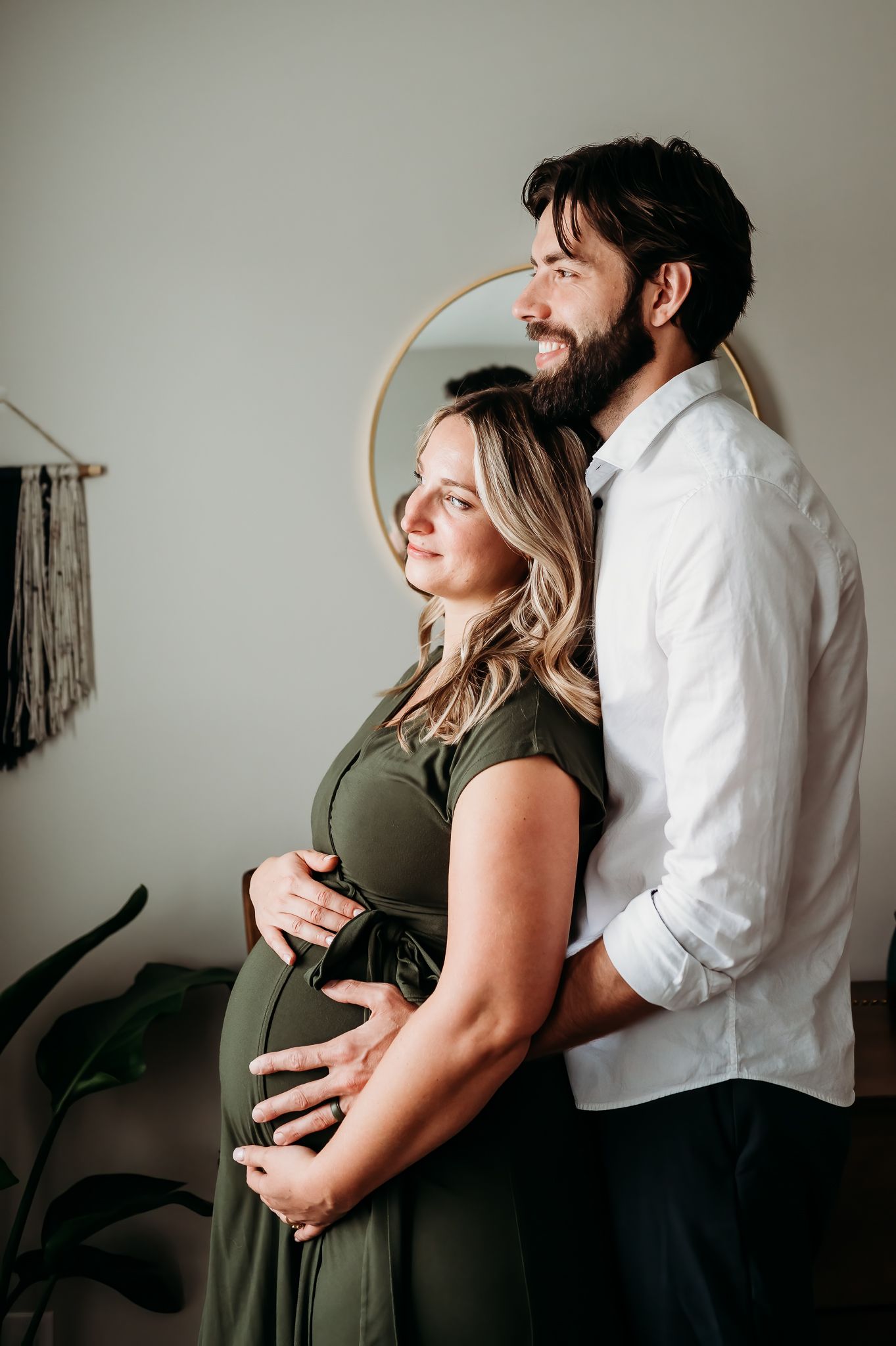 Expecting couple embraces in a tender maternity photo with olive green dress and white shirt against neutral wall.
