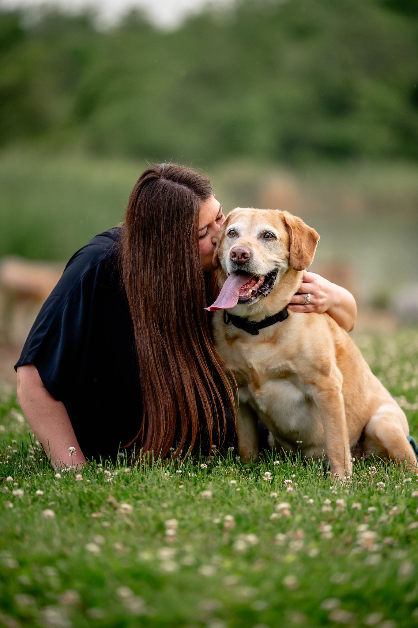 Young woman with long brown hair kissing her golden Labrador Retriever while sitting on green grass outdoors.