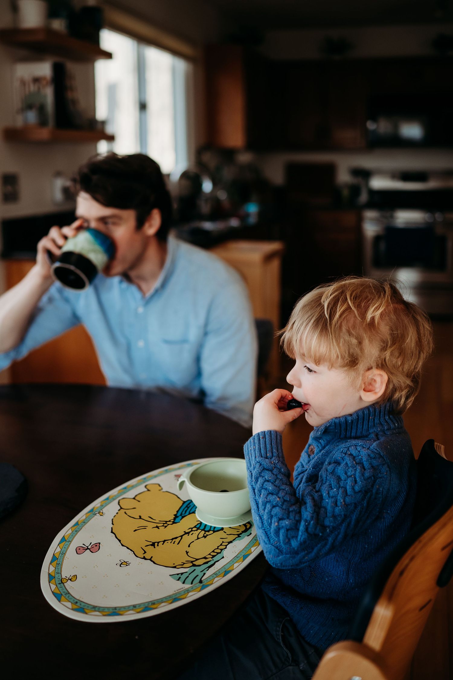 A toddler in a blue sweater eats at a table with an illustrated plate while a man drinks from a mug nearby.