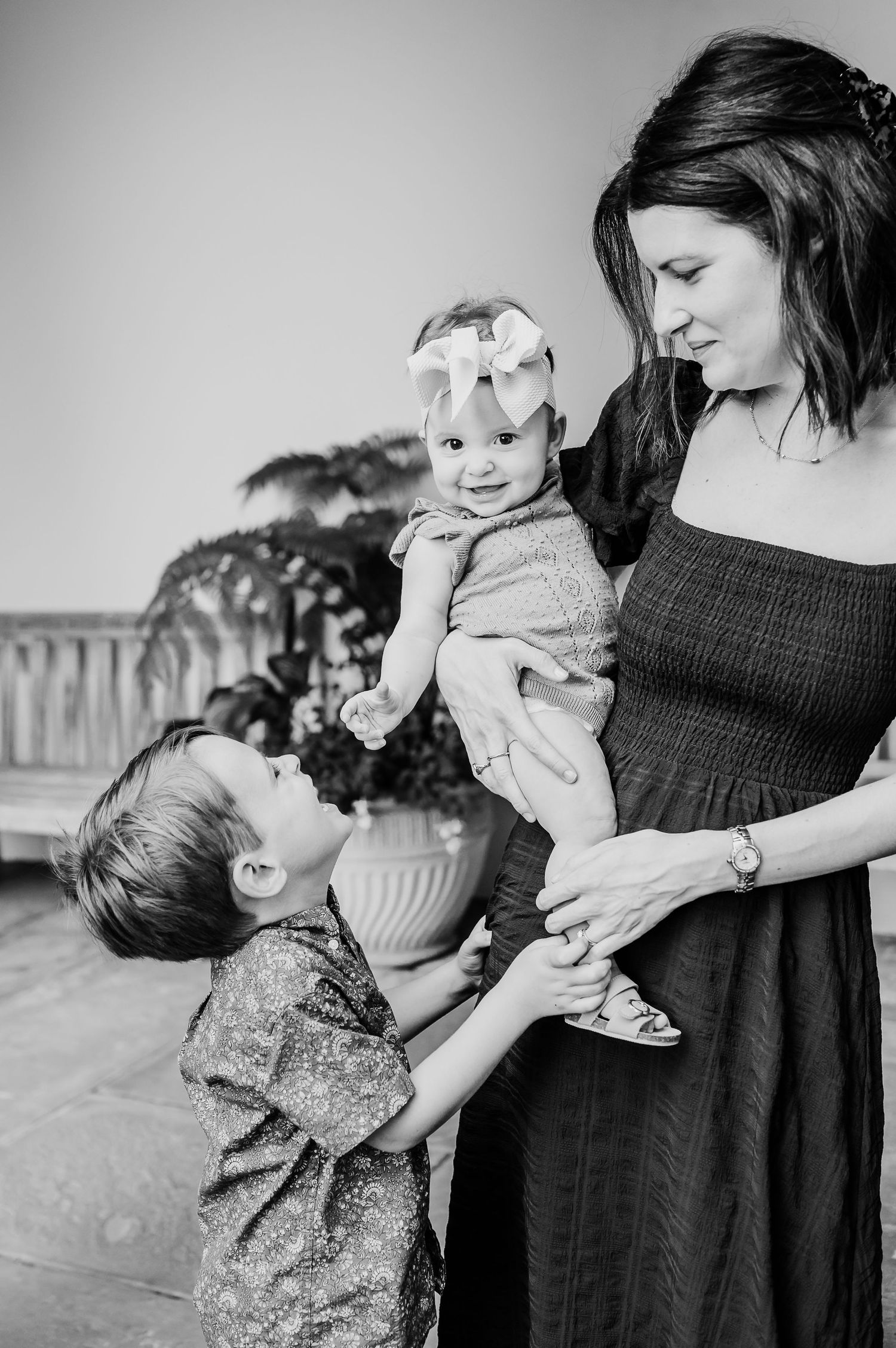 Black and white photo shows siblings sharing playful moment as mom smiles at them.