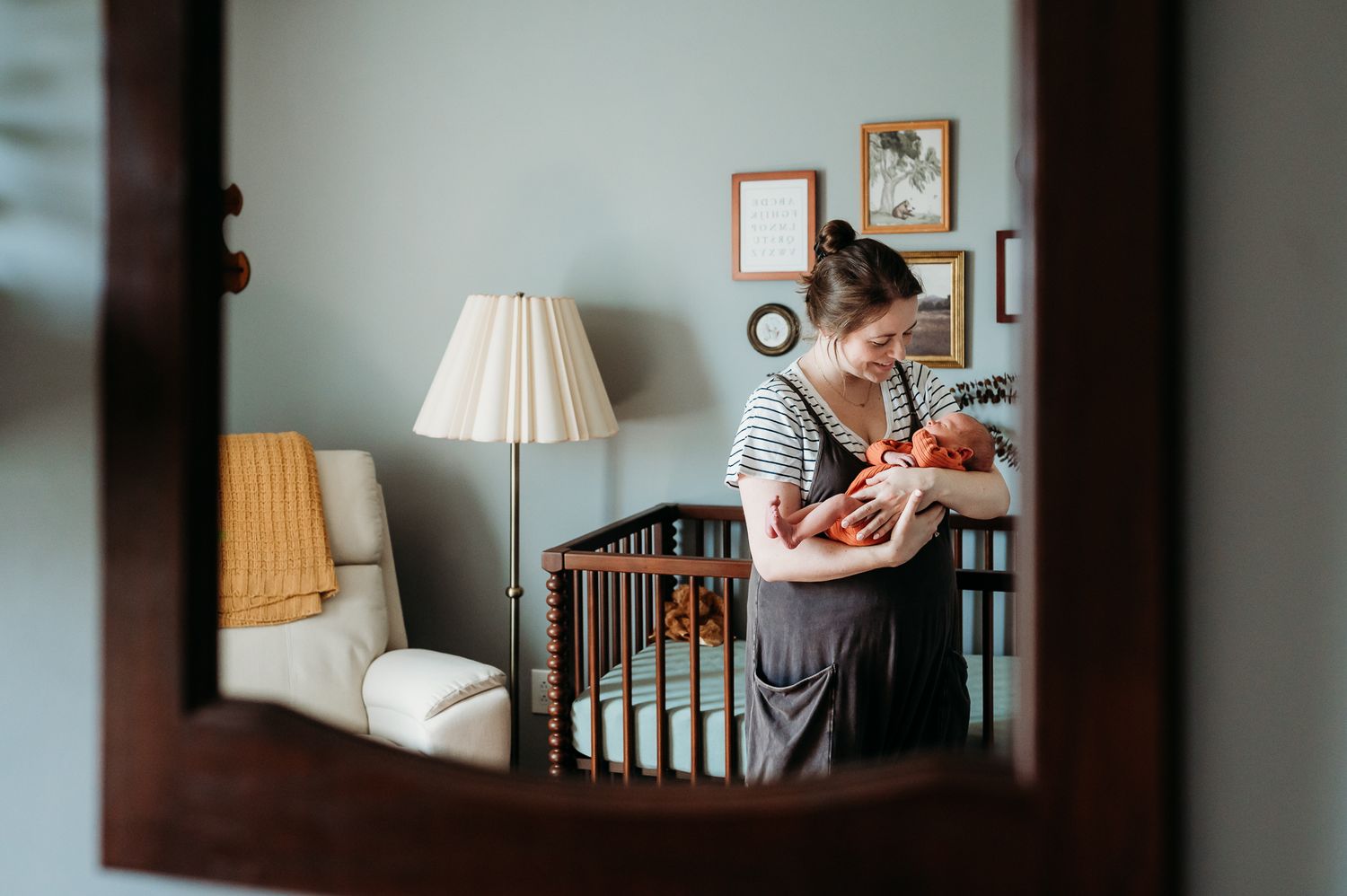 Reflection in mirror shows mother holding baby in striped shirt next to wooden crib in nursery during photo session in Indiana home.