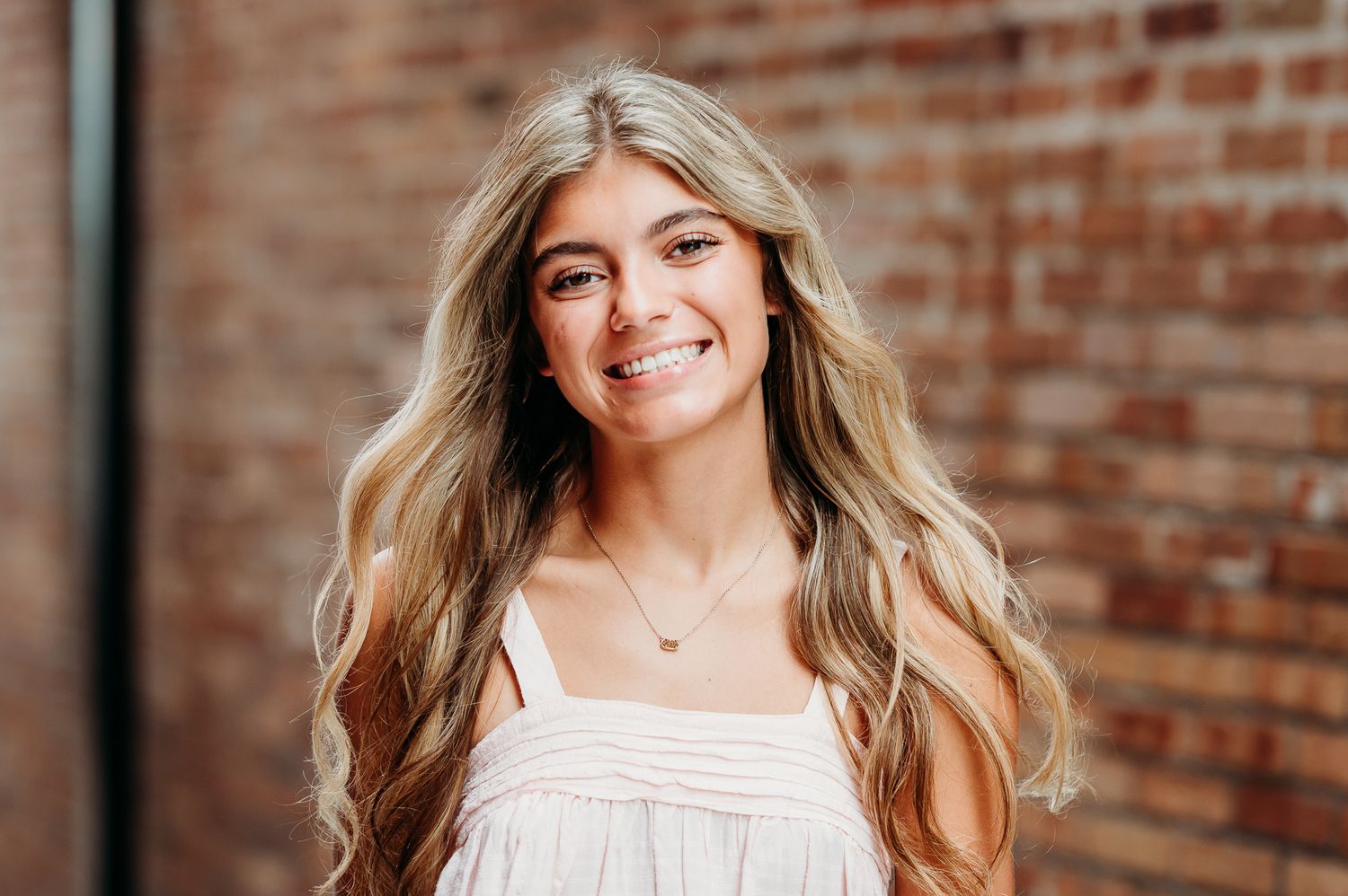 A person with long wavy blonde hair poses outdoors against a rustic brick wall while wearing a white summer dress.