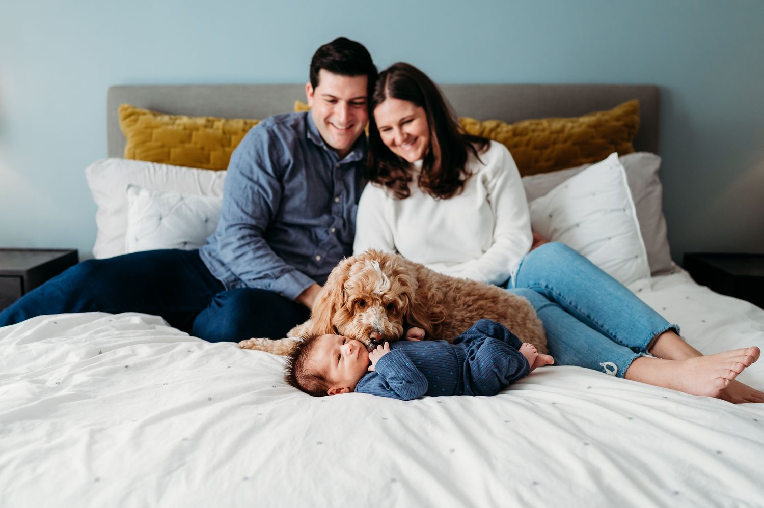 A couple relaxes on a white bed with their goldendoodle puppy in a cozy bedroom setting.
