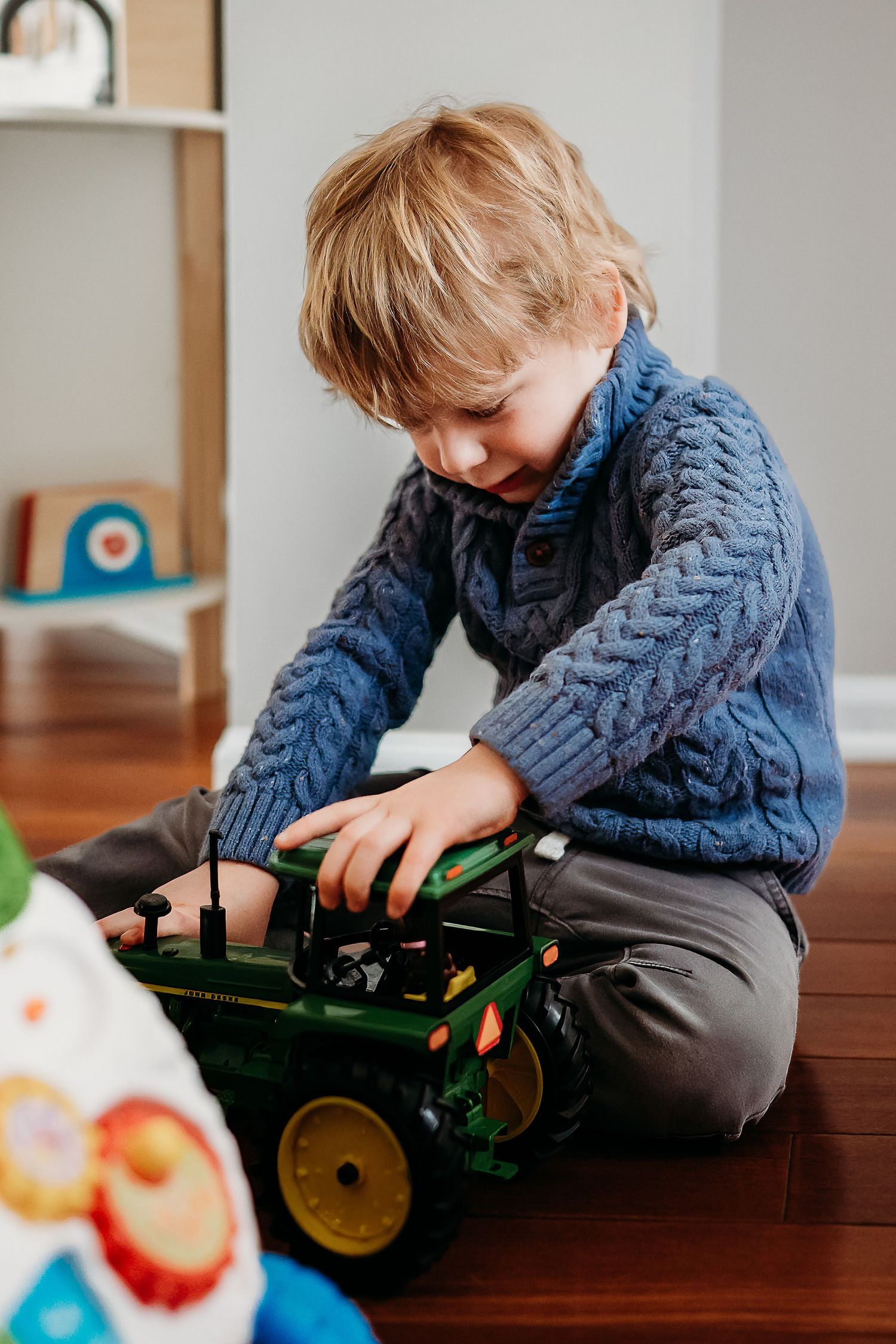 Young boy in blue cable-knit sweater playing with a green toy tractor on hardwood floor.
