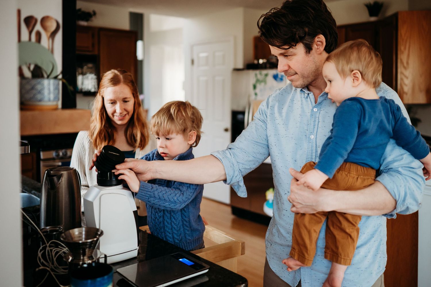 Family with two young children making coffee together in kitchen, parents smiling as toddler uses appliance.