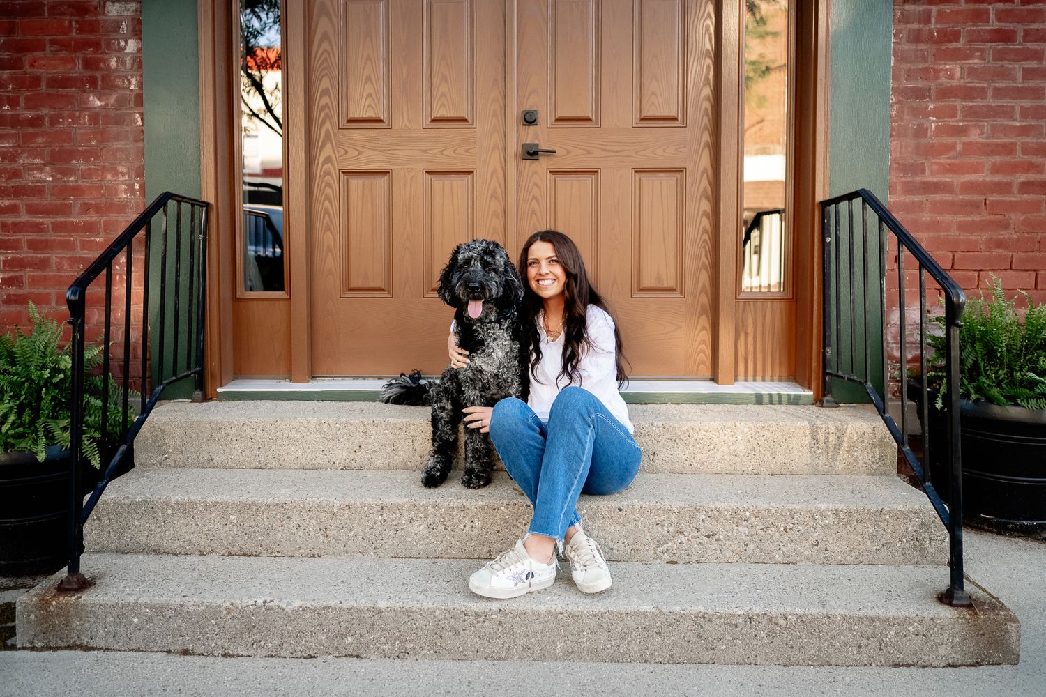 A young woman sits smiling on brick building steps with a black dog beside her, wearing jeans and white sneakers.