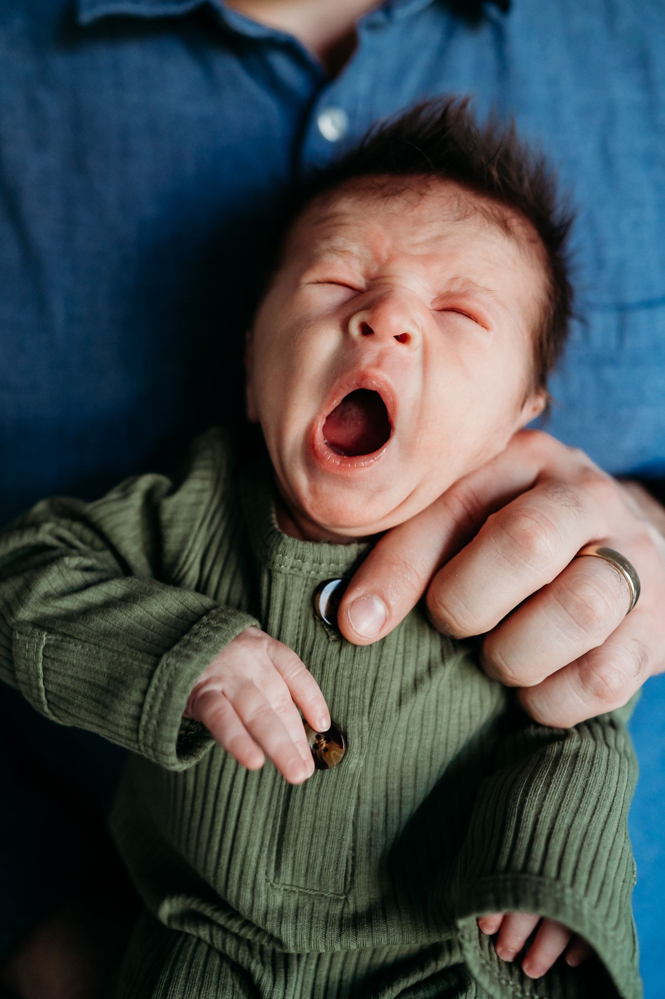 Baby in green ribbed sweater yawning widely while being held against blue background.