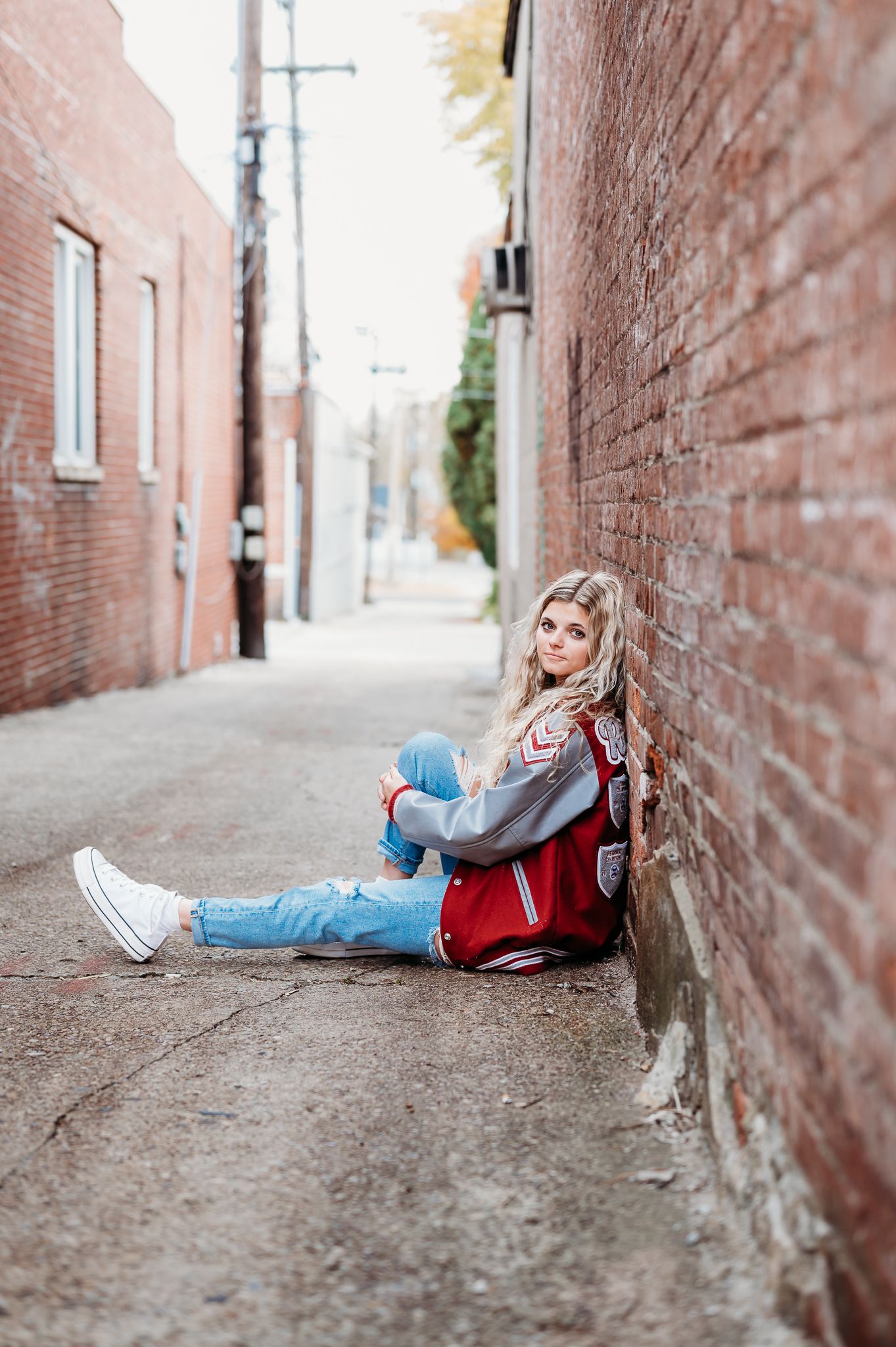A senior girl sits casually against a brick wall in an urban alley wearing a red jacket and jeans.