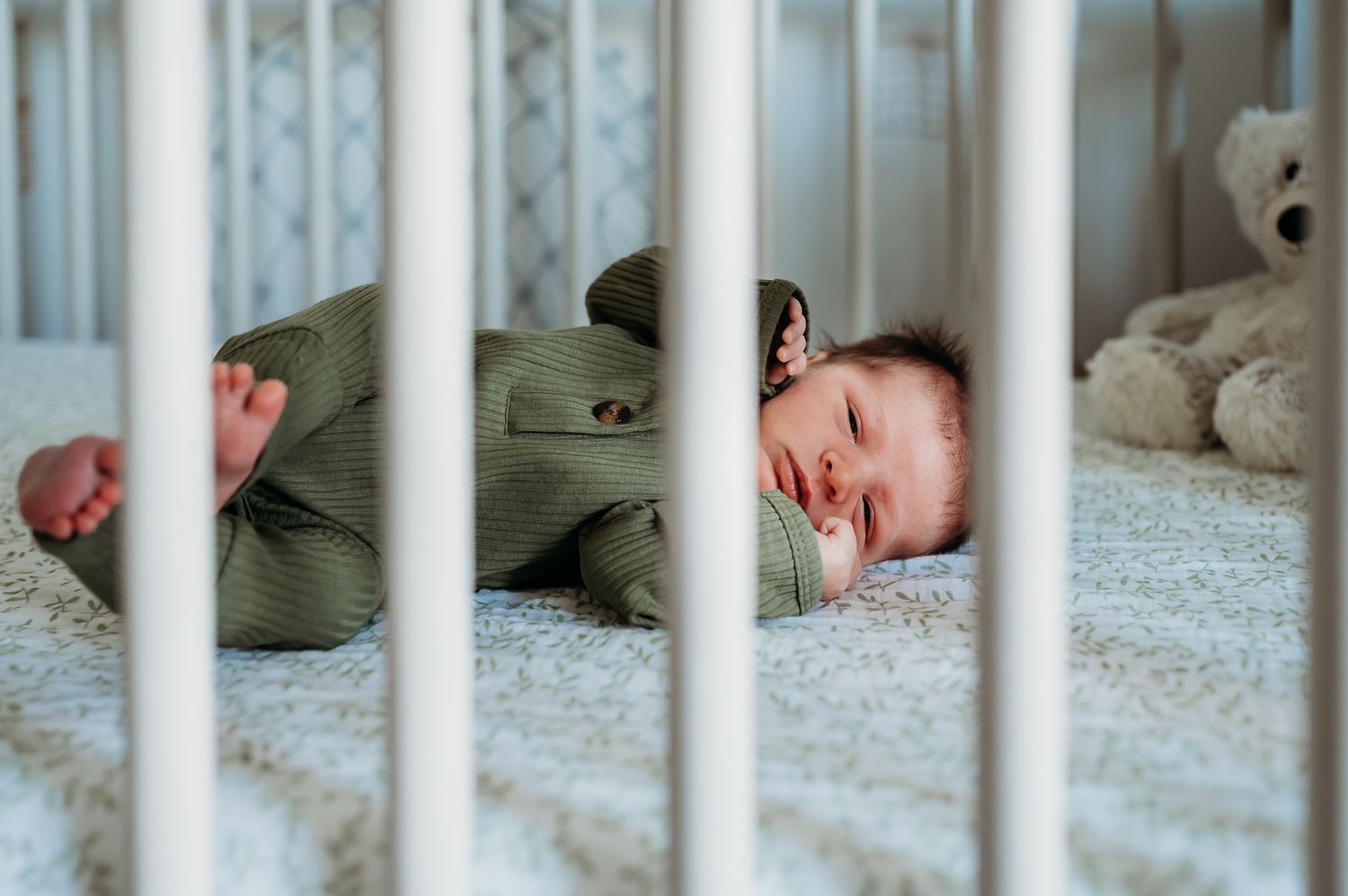 A sleeping baby in a green onesie rests peacefully inside a white crib with a teddy bear nearby.