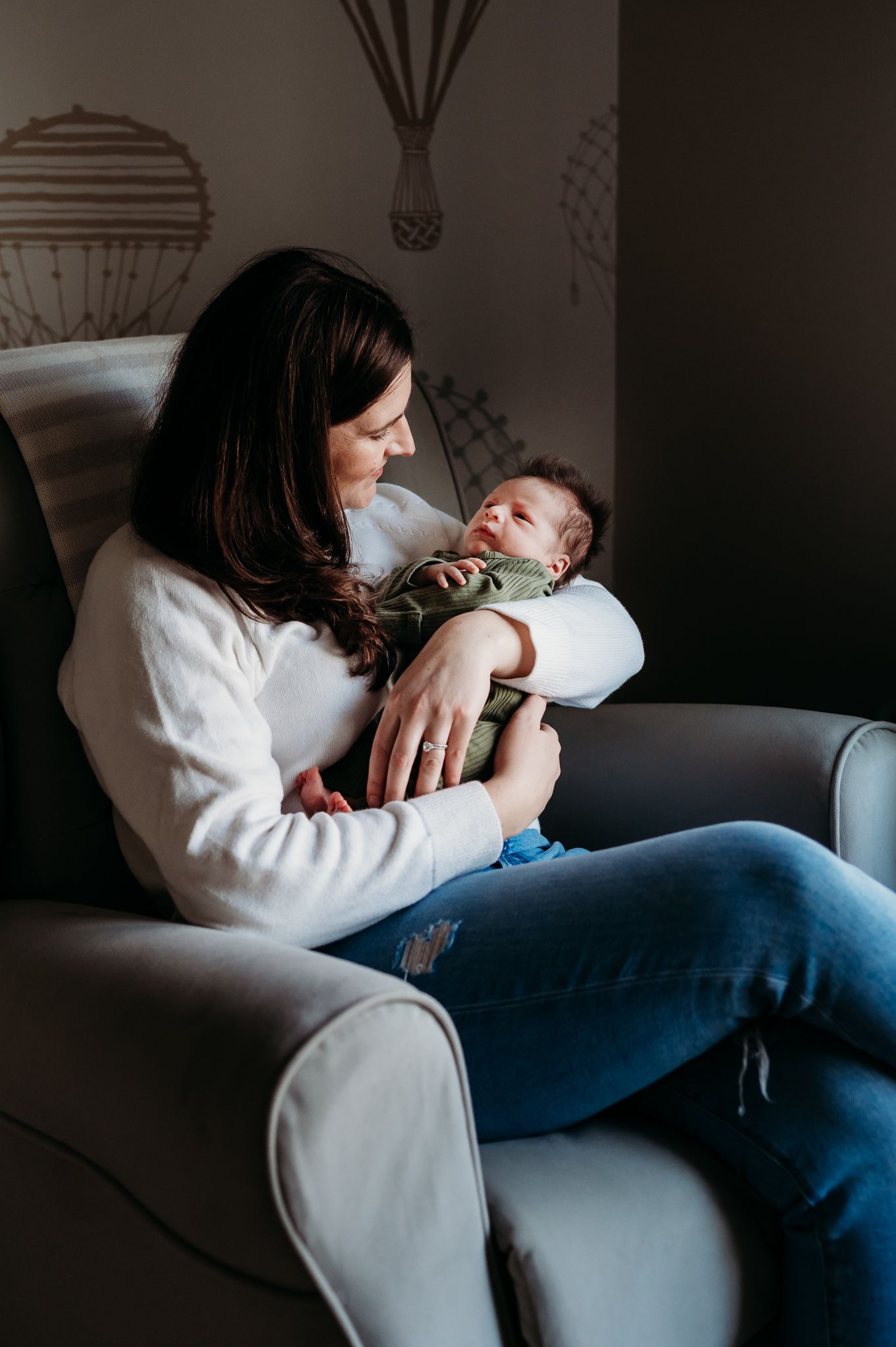 Mother in a white sweater and jeans sitting in a gray armchair holding her small baby in a heartwarming moment.