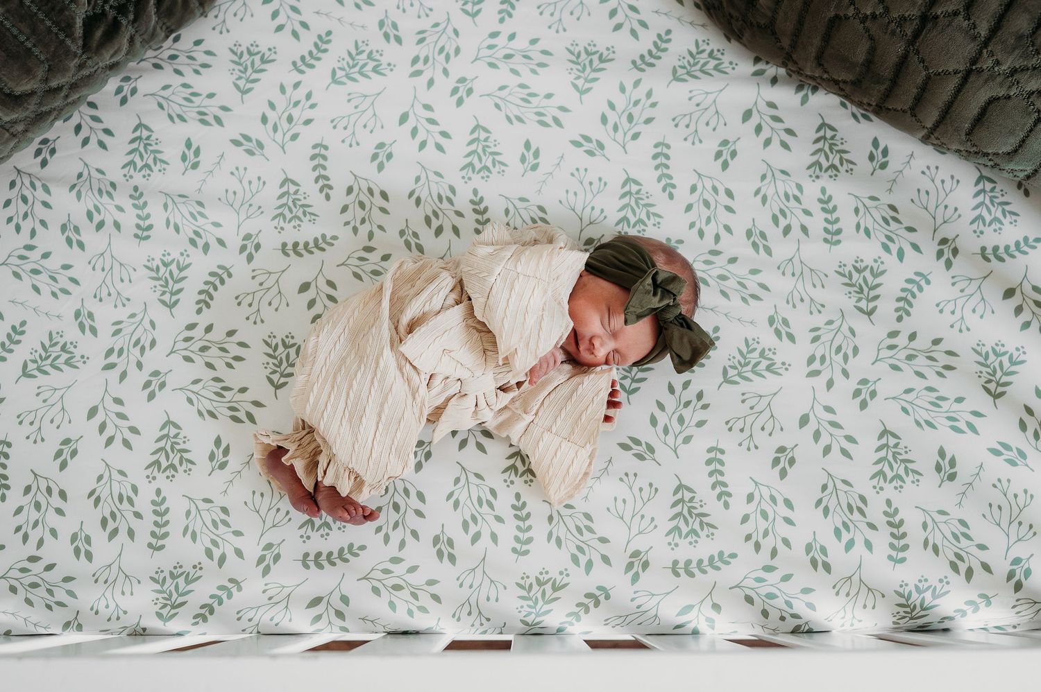 Newborn baby swaddled in cream wrap sleeping peacefully in a crib with white floral leaf print sheets.