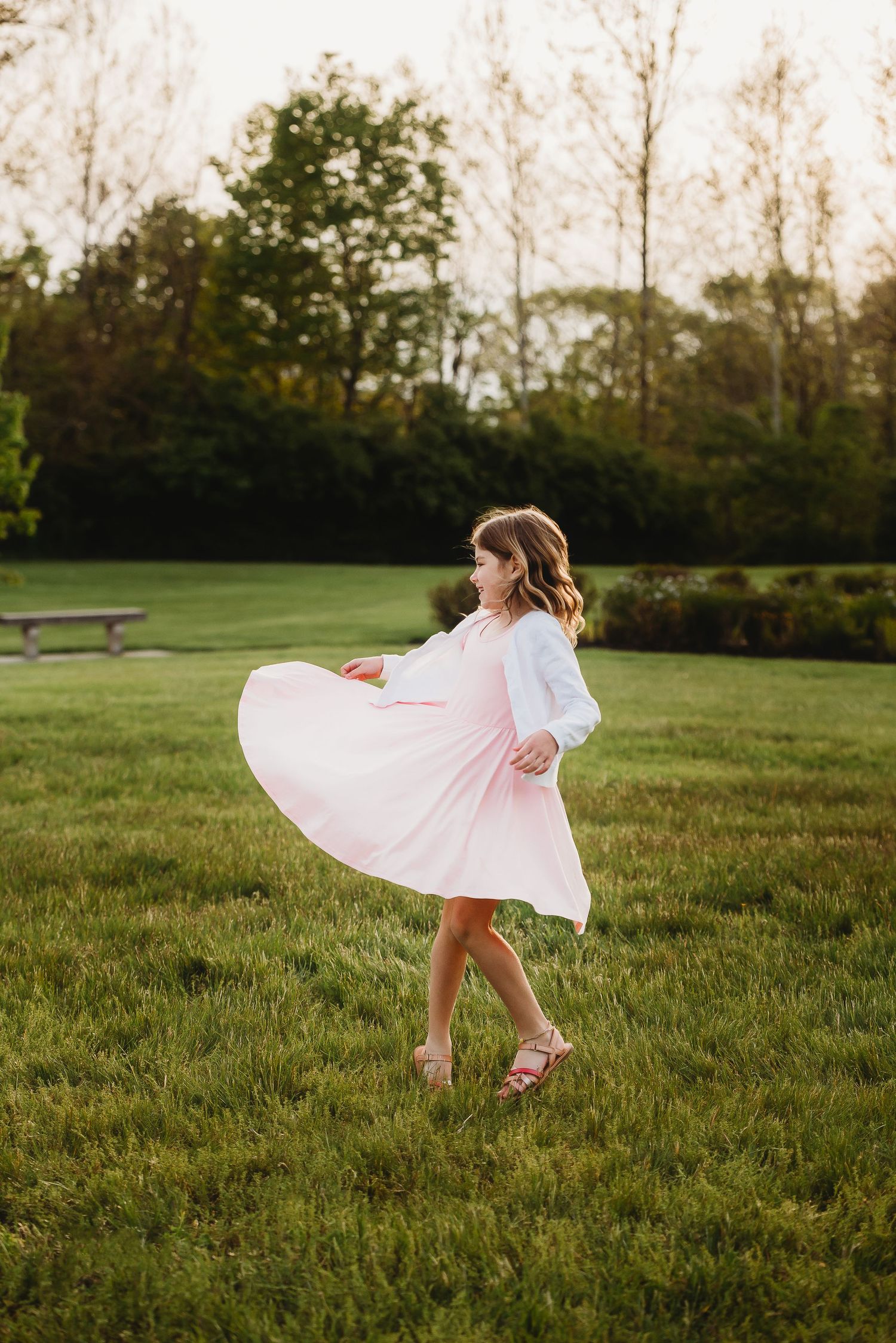 Little girl in pink dress twirls playfully in a sunlit meadow during spring family session at Friendship Gardens in Plainfield, Indiana