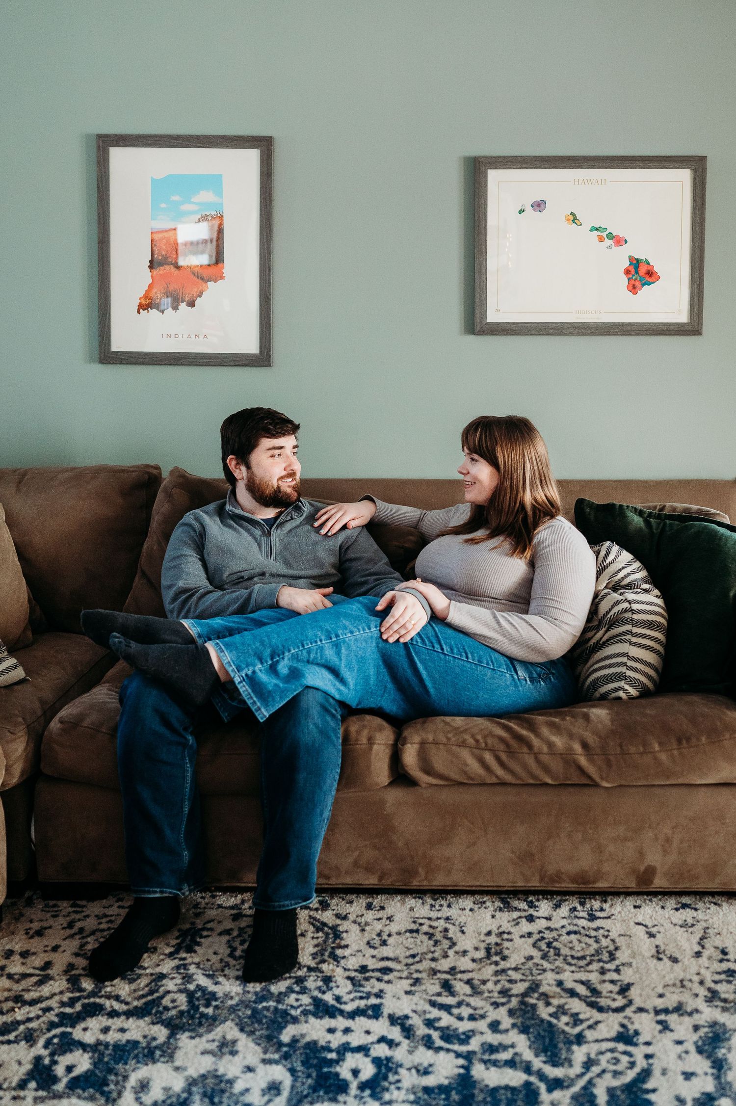 A couple relaxes on a brown couch, chatting warmly with framed maps on a sage green wall during in-home Indianapolis couples photoshoot.