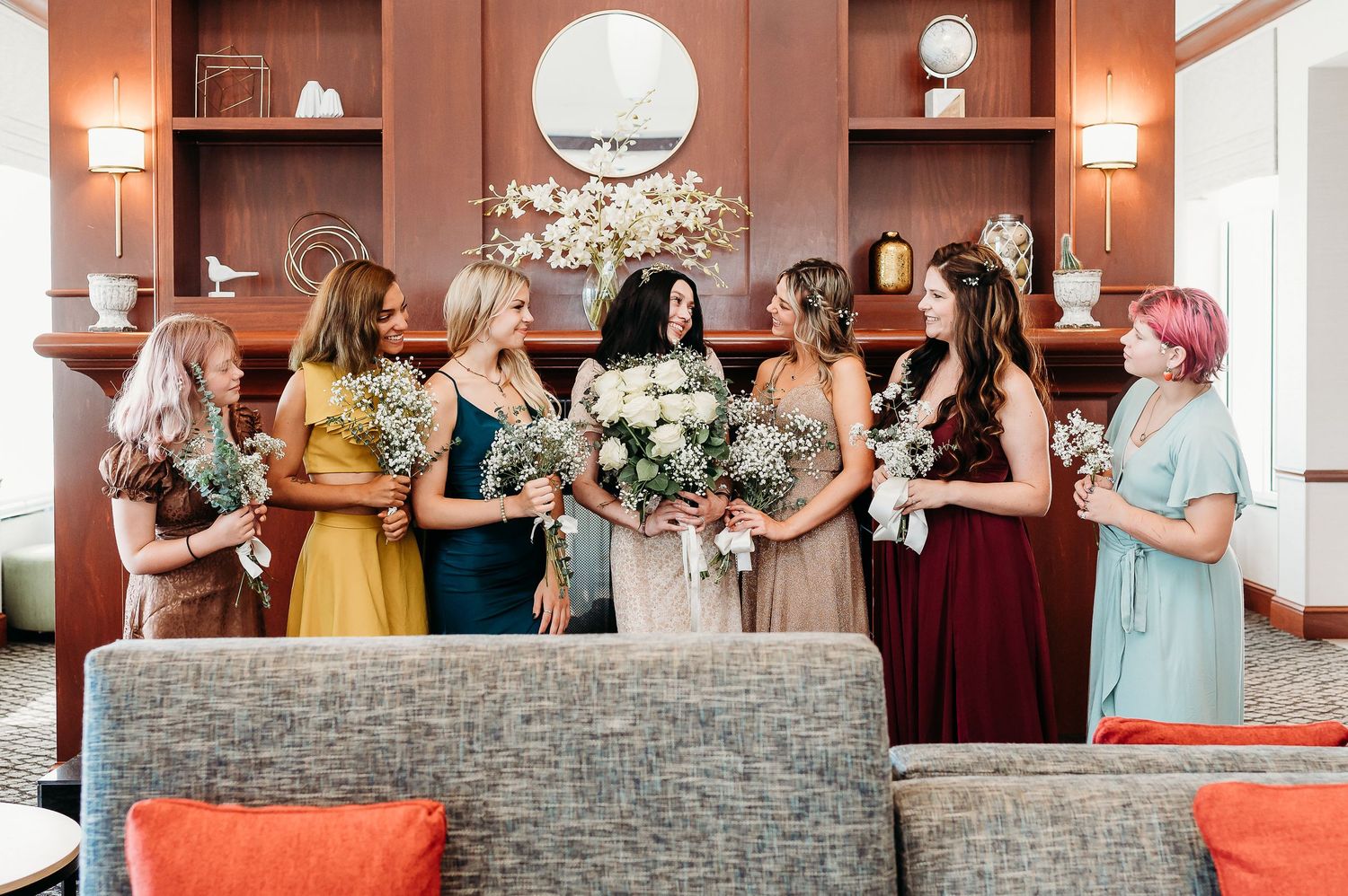 Wedding party in mix of burgundy and yellow dresses gather around couch with floral bouquets.