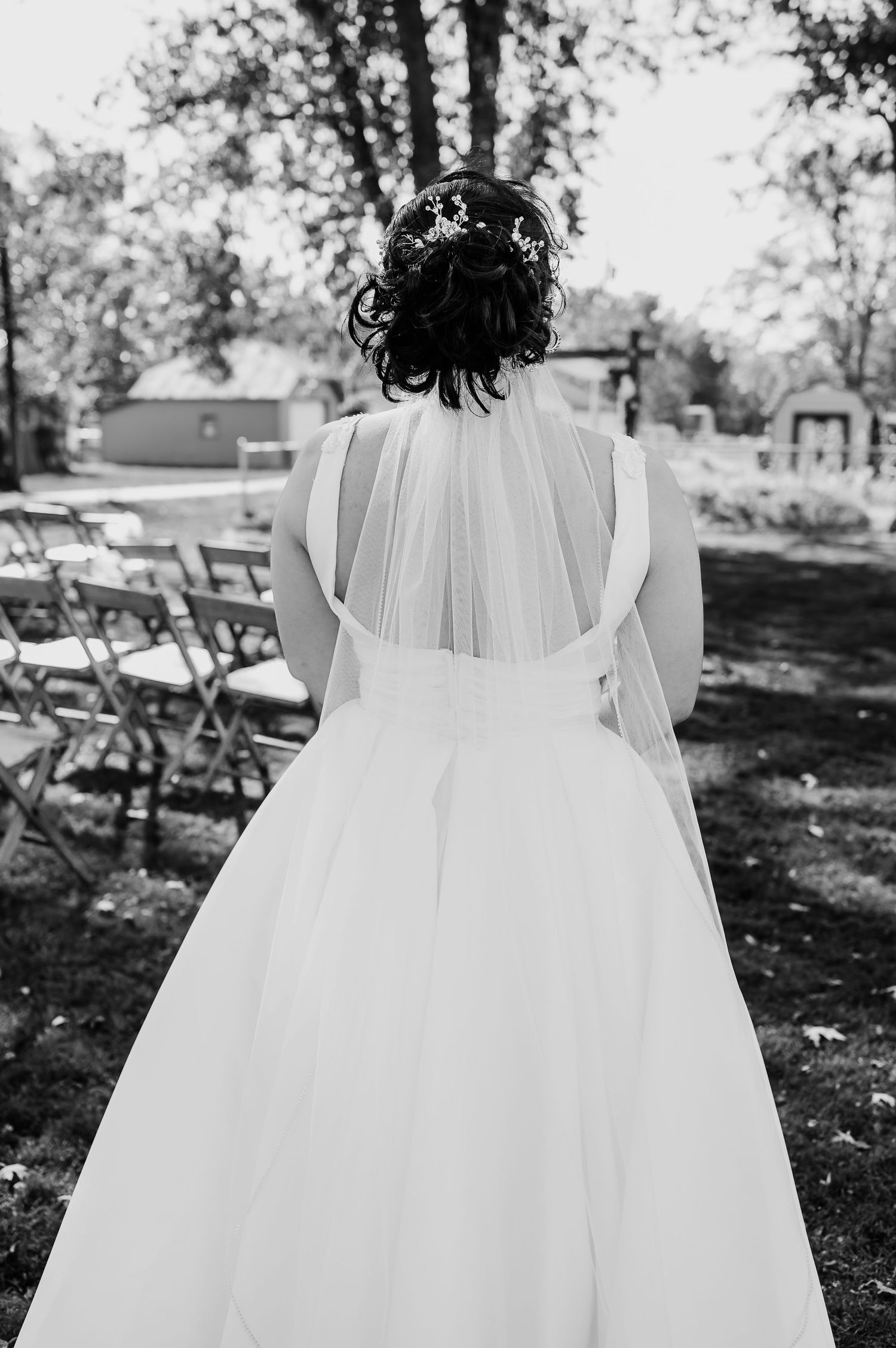 Black and white photo of bride in flowing white wedding dress with sheer back detail standing in outdoor garden setting.