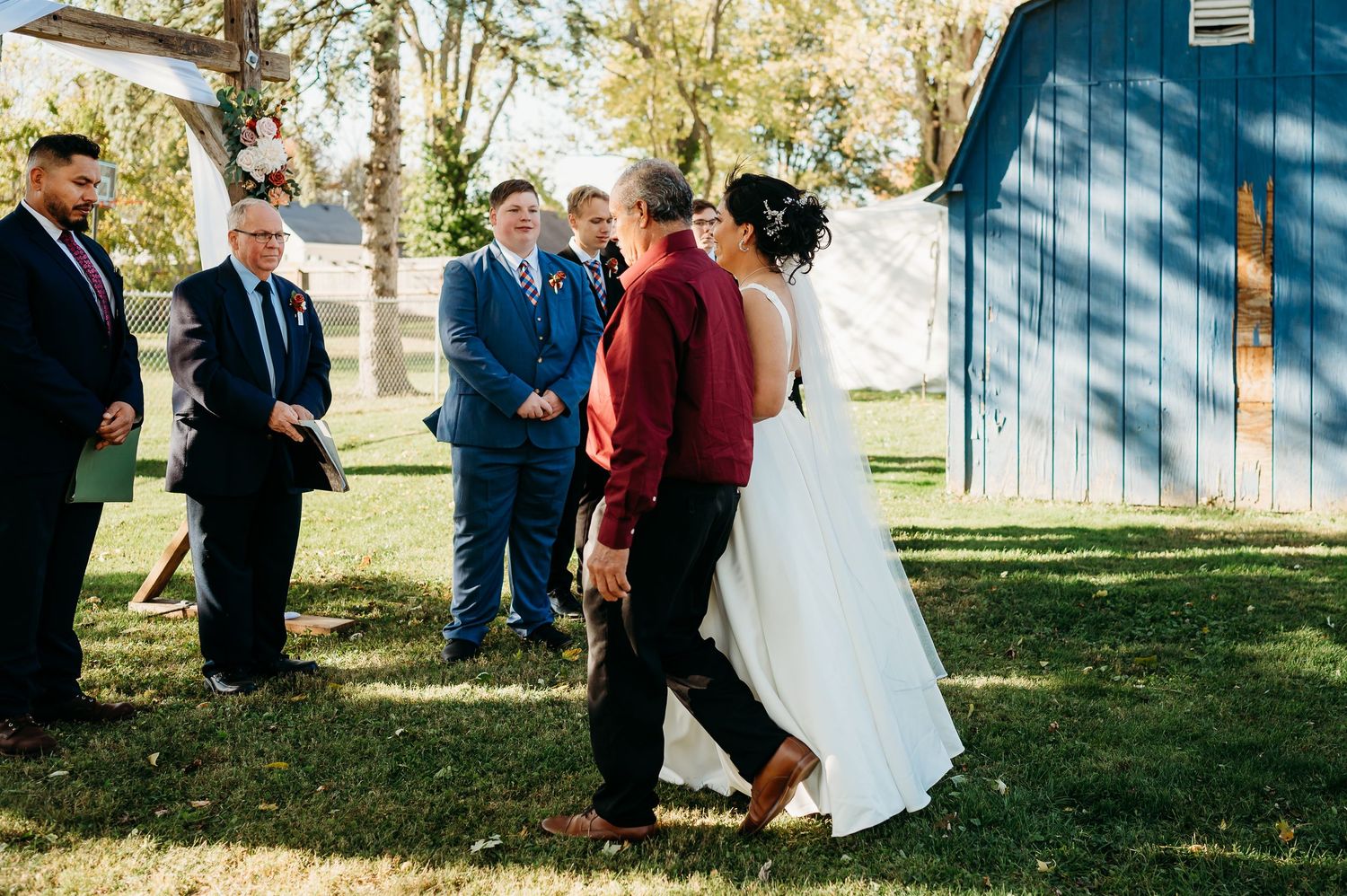 Wedding party stands in line against blue barn wall with sunlit trees in background.