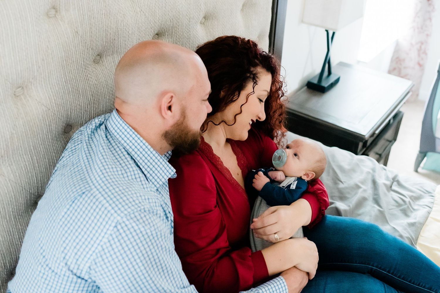 A couple snuggles together on bed, tenderly looking down at their newborn baby wearing blue outfit during newborn photos at Avon, IN, home.
