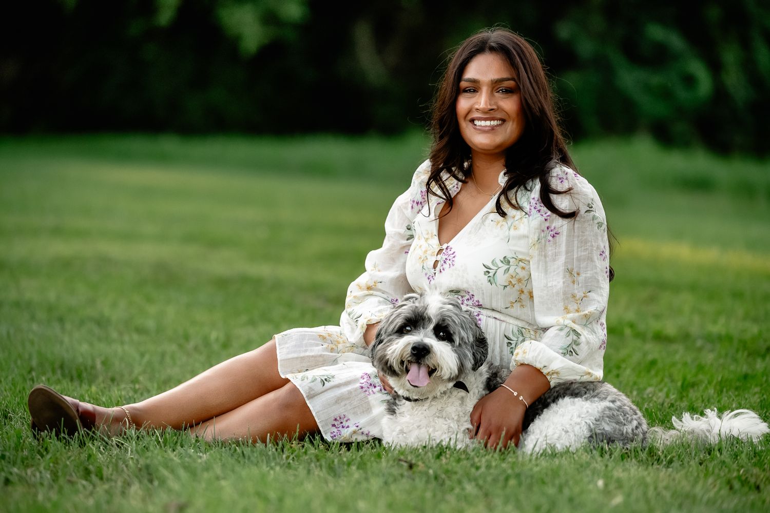 Young woman in floral dress sitting on grass smiling with her gray and white mini bernesdoodle during dog photo session in Plainfield, IN.
