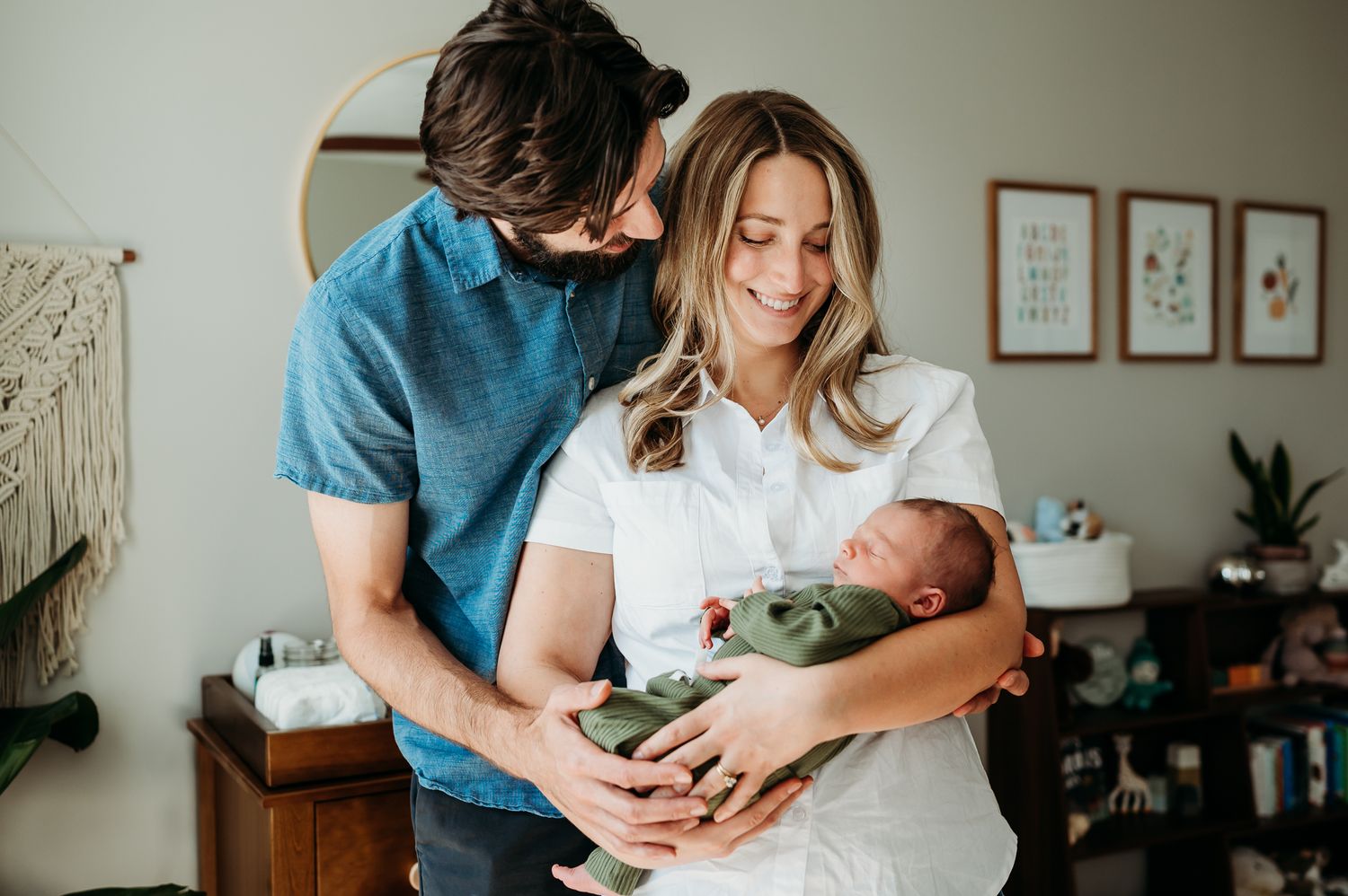 Parents share a tender moment with their newborn baby in a cozy nursery with macrame wall art and plants.