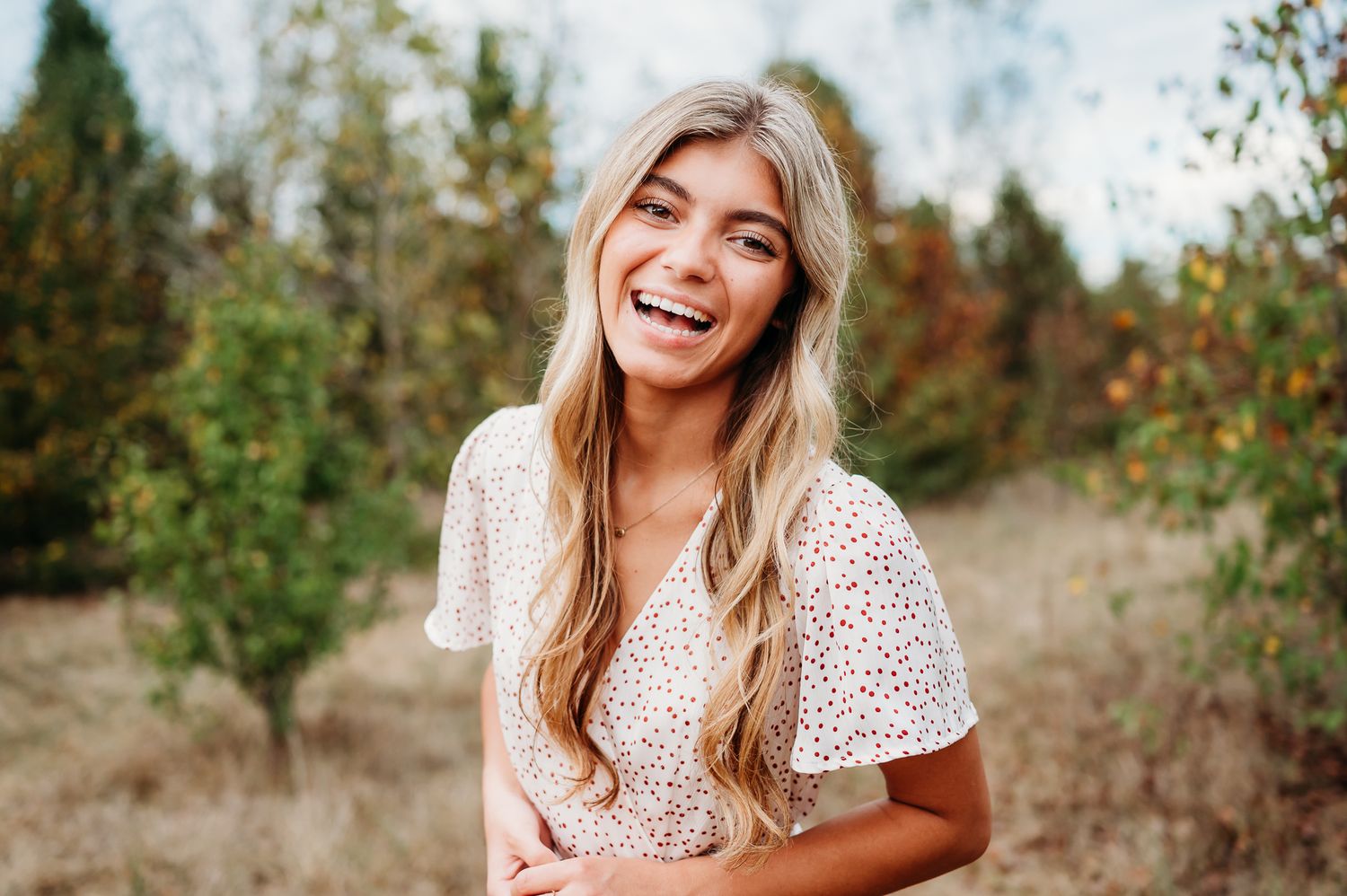 Someone in a white eyelet dress laughs joyfully outdoors with a natural backdrop of trees and autumn foliage.