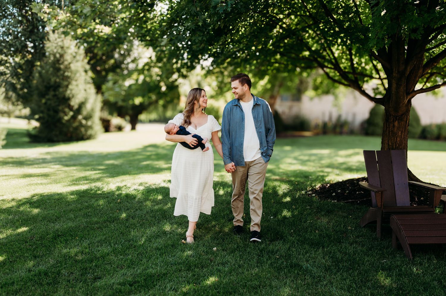 A couple walks together on a sunny day in a lush Carmel, Indiana, garden setting holding baby and smiling.