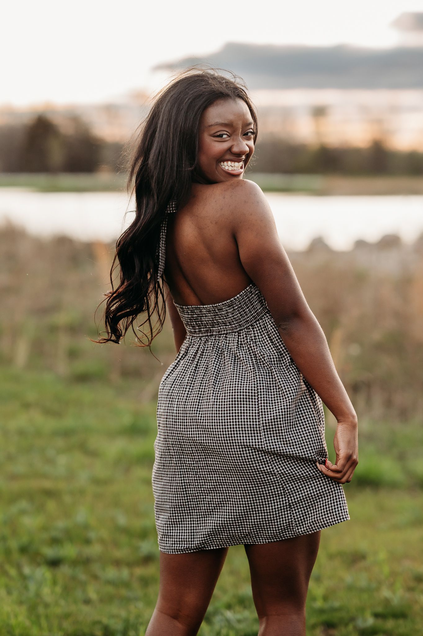 A smiling woman in a checkered sundress poses outdoors at sunset near a lake with long flowing dark hair.