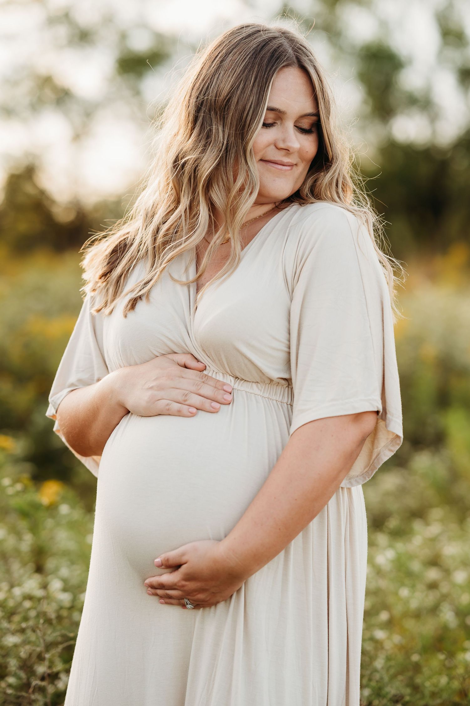 A maternity photo shows pregnant woman in cream dress cradling their baby bump in a sunlit wildflower field during Indy maternity photoshoot