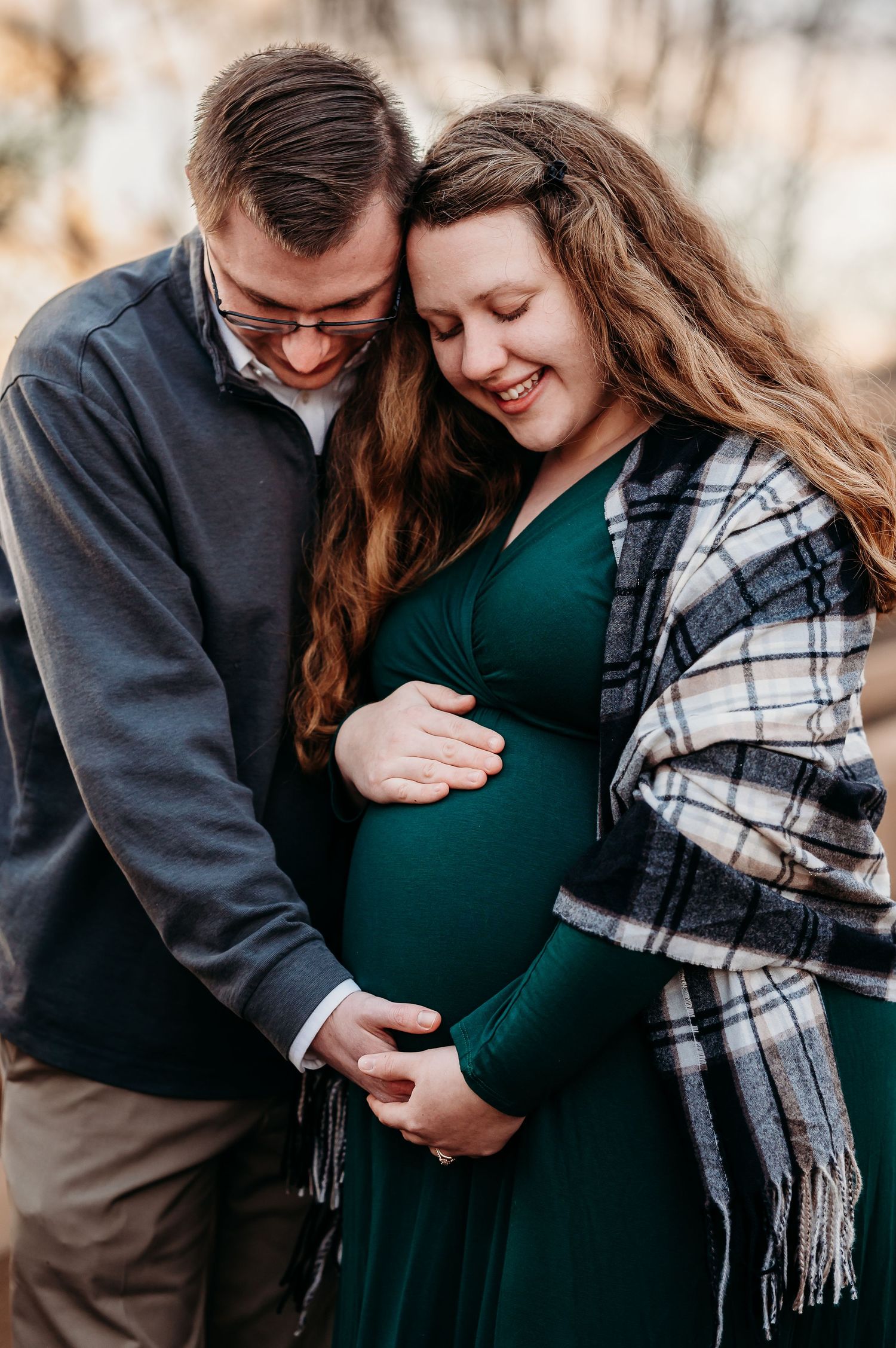 A joyful pregnant couple embraces outdoors, woman in green dress with plaid shawl, both cradling her baby bump.