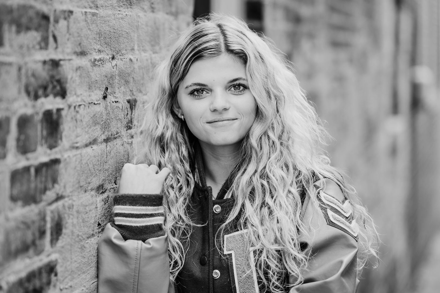 Black and white portrait of a person with long wavy blonde hair wearing a varsity jacket and posing against a brick wall.