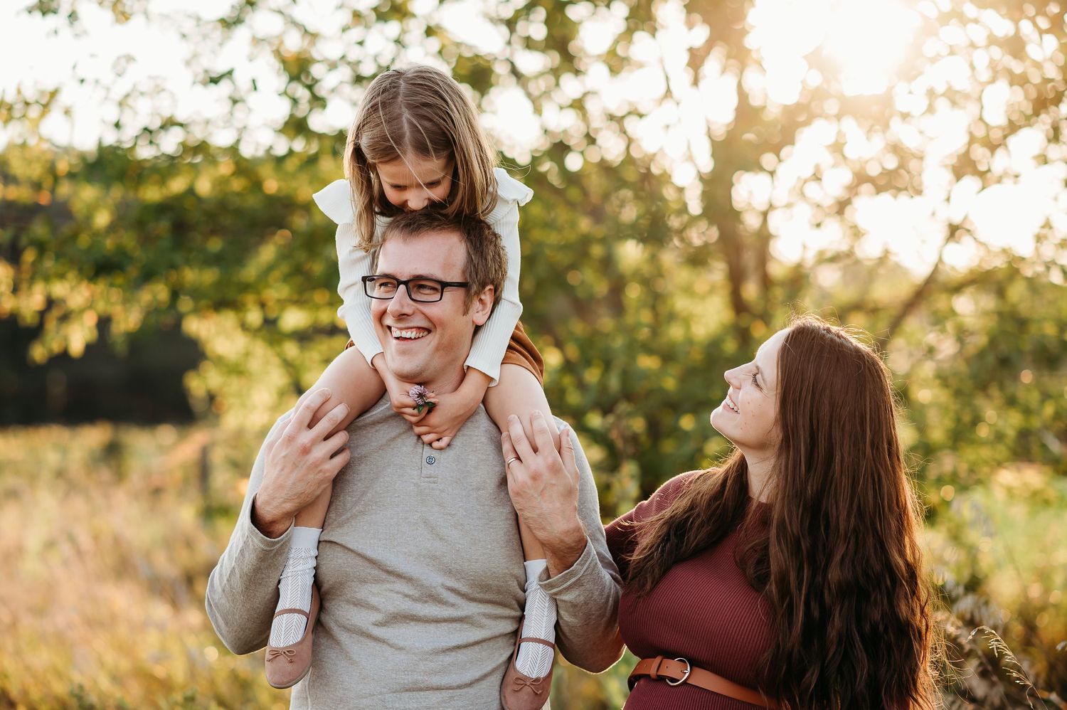 Couple and daughter embraces in a sunlit field in West Lafayette during golden hour while laughing and sharing a joyful moment together.