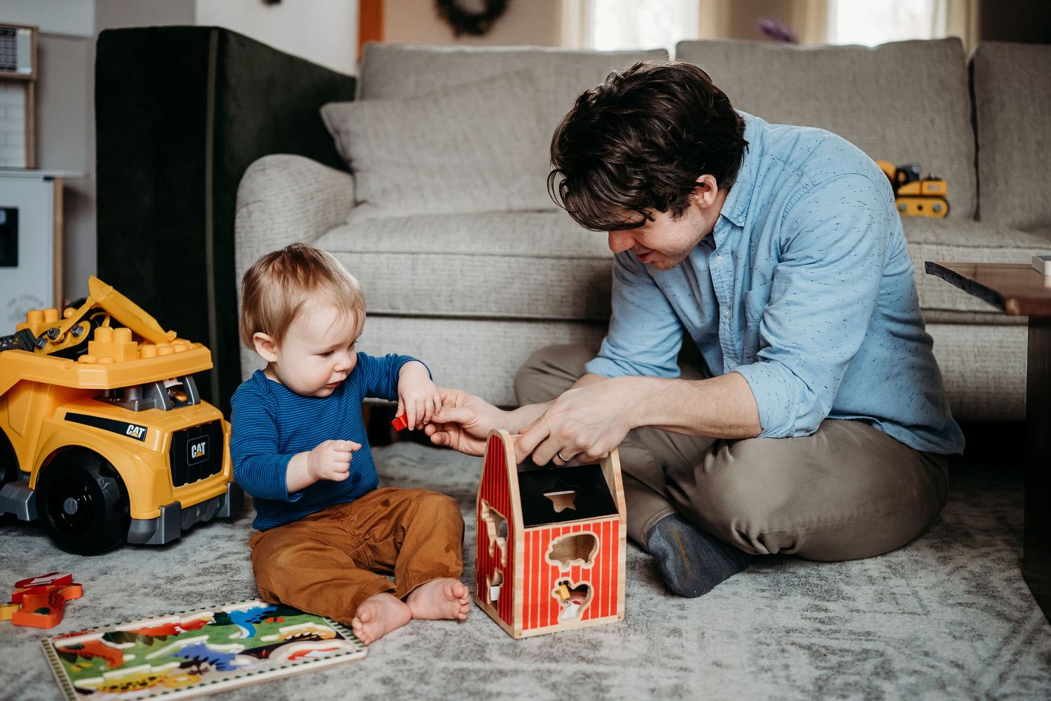 Father and toddler playing together on living room floor with wooden toy barn and colorful children's toys.