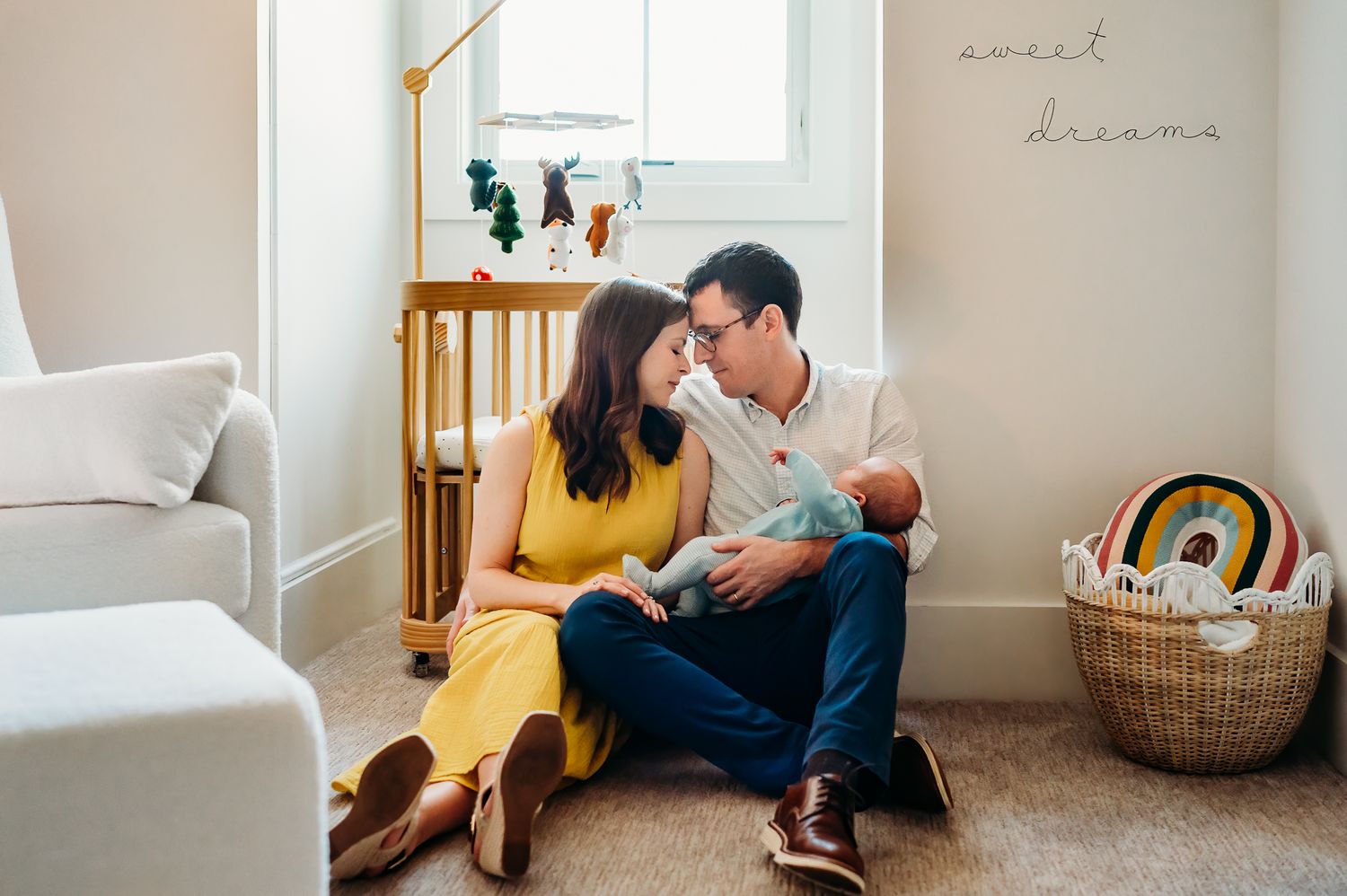 A Westfield couple sits on the floor of a cozy nursery with a rainbow toy basket nearby as they share cuddle baby boy.